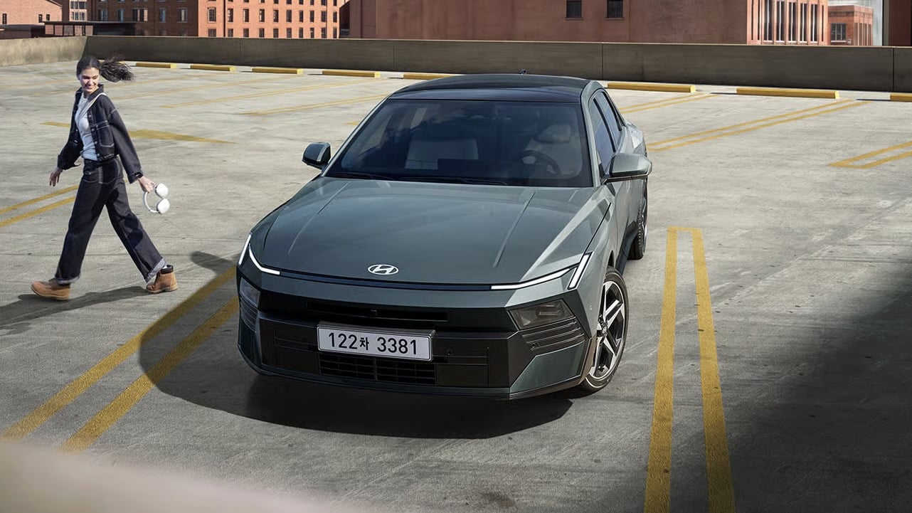 Gray Hyundai sedan parked on a rooftop parking area with yellow lines and city buildings in the background; a person walks past holding white objects in their hand and hair in motion.