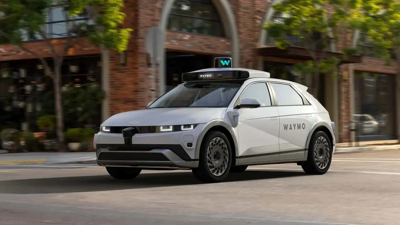 White Waymo self-driving SUV with rooftop sensor pod driving on a city street with brick buildings in the background and Waymo branding on the side.