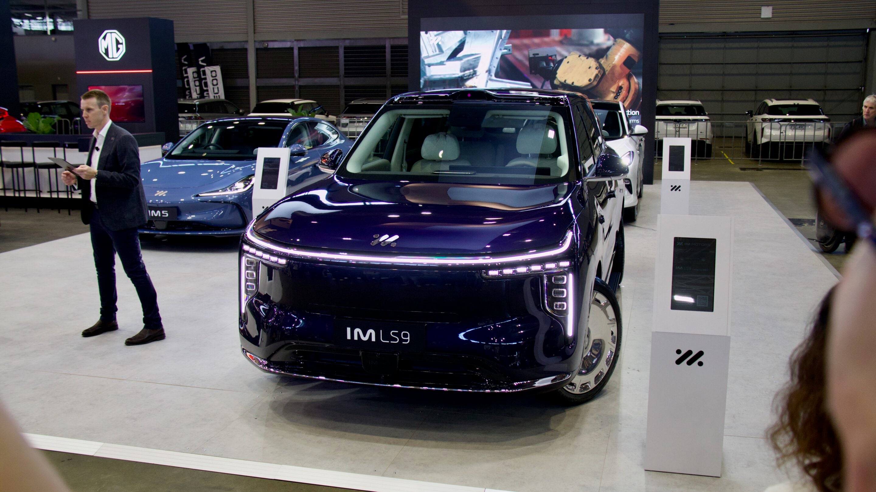 A man in a blazer and jeans stands beside a glossy dark blue SUV at a car exhibition, with other cars on display in the background.