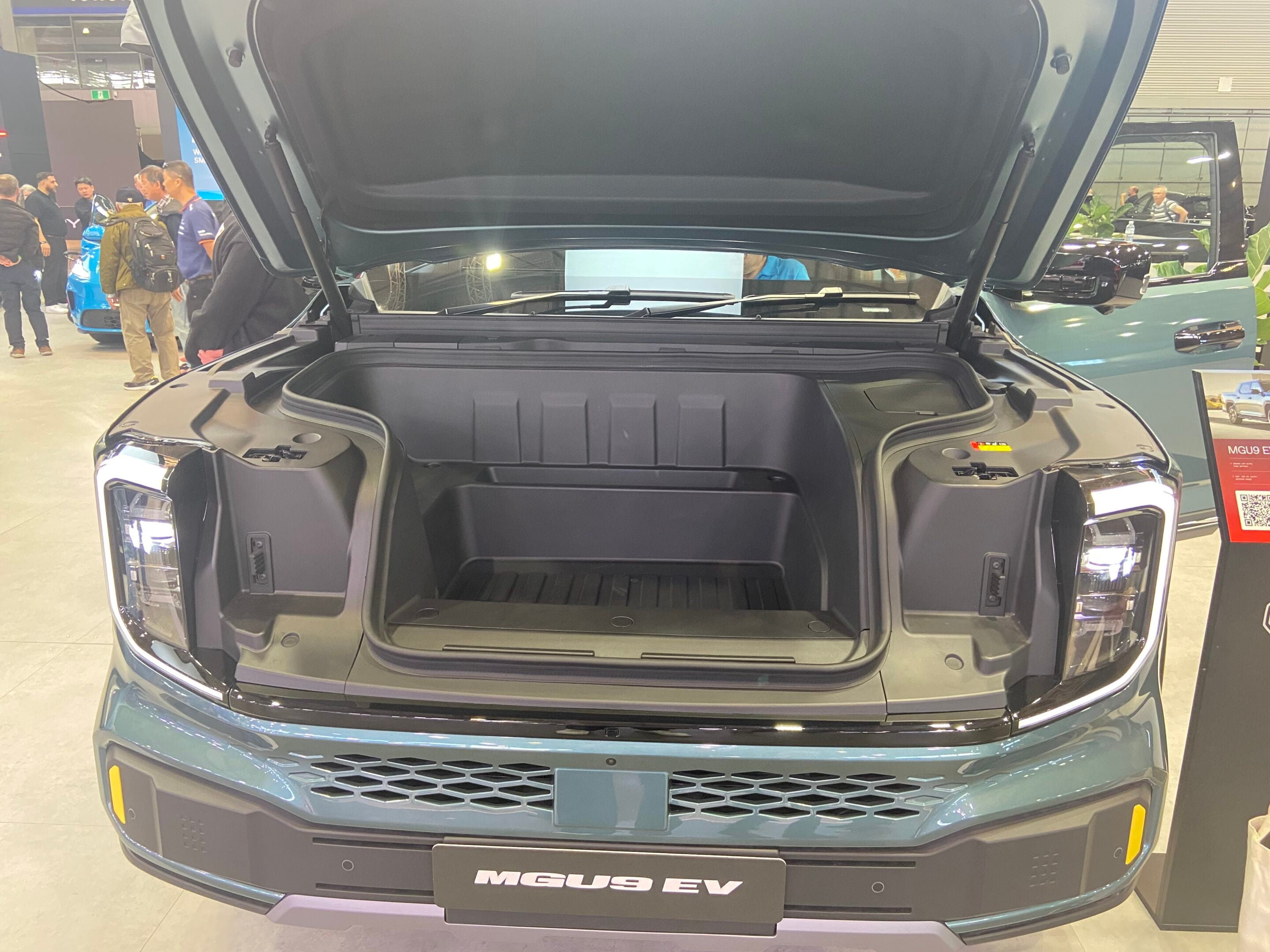 Front view of an electric SUV with its trunk open at an auto show, MG EV badge visible on the front plate area.
