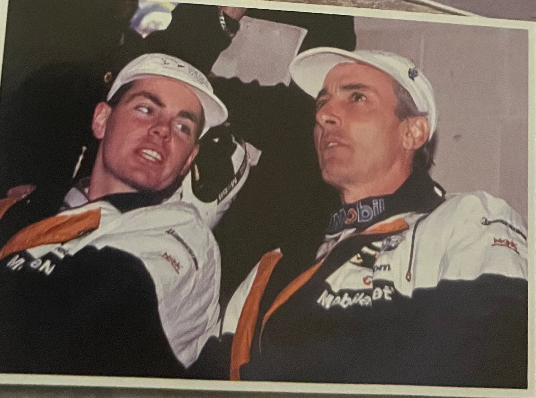 Two men in white racing suits with orange lanyards pose together, wearing white caps, likely after a race.