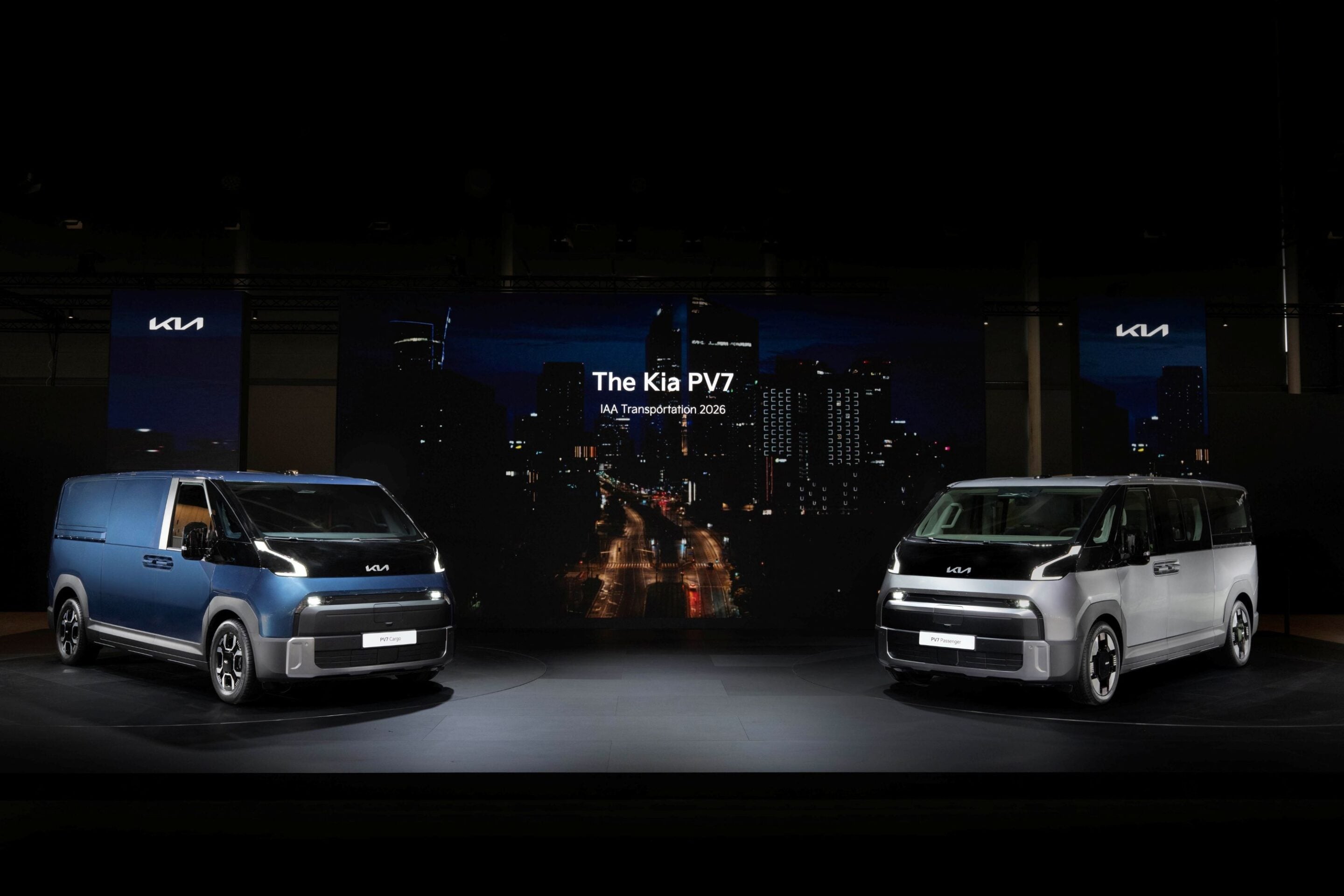 Two Kia PV7 vans on a showroom floor with a cityscape screen in the background displaying 'The Kia PV7'.