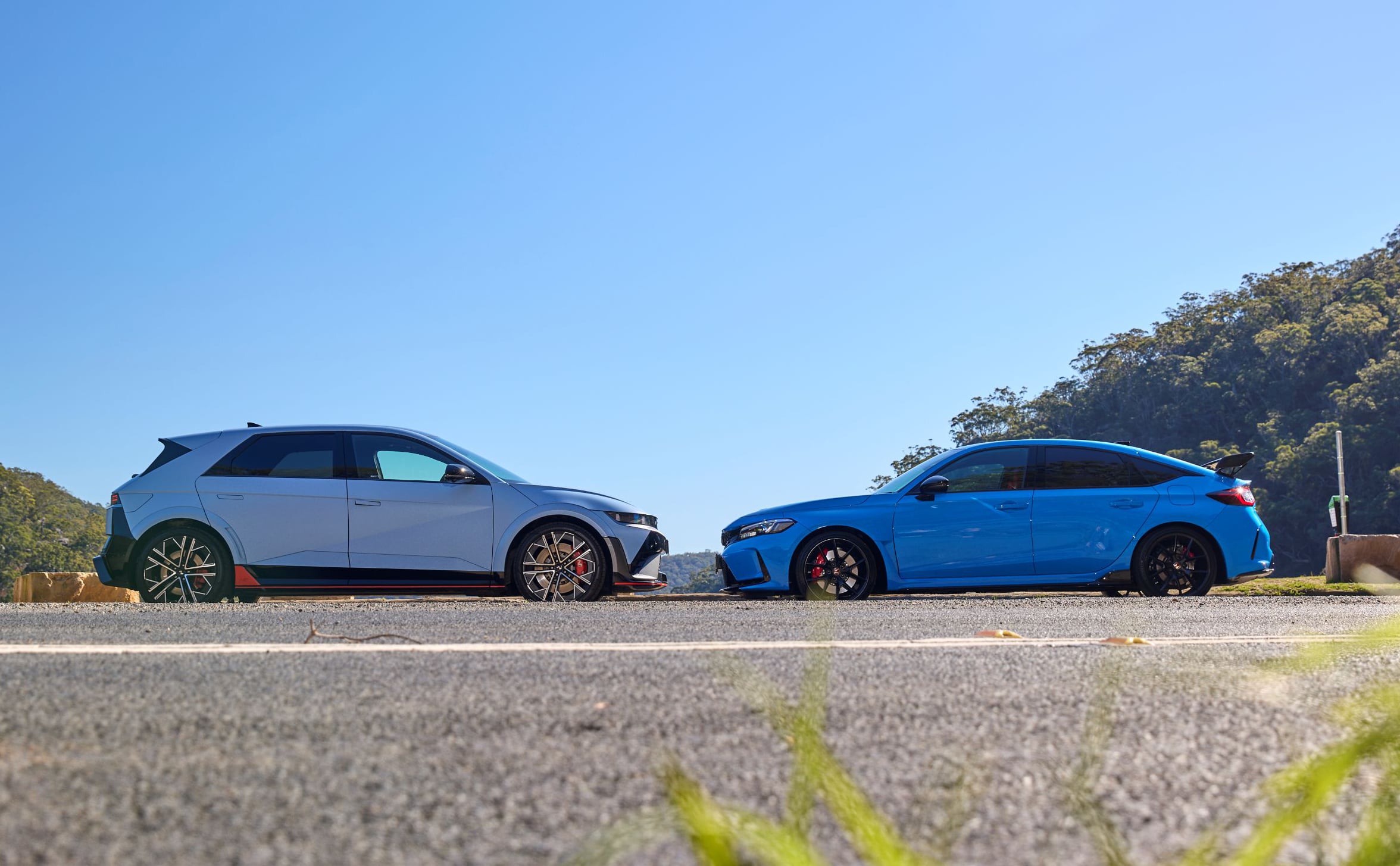 Two sporty cars parked nose-to-nose on a sunny road with a clear blue sky and green hills in the background.