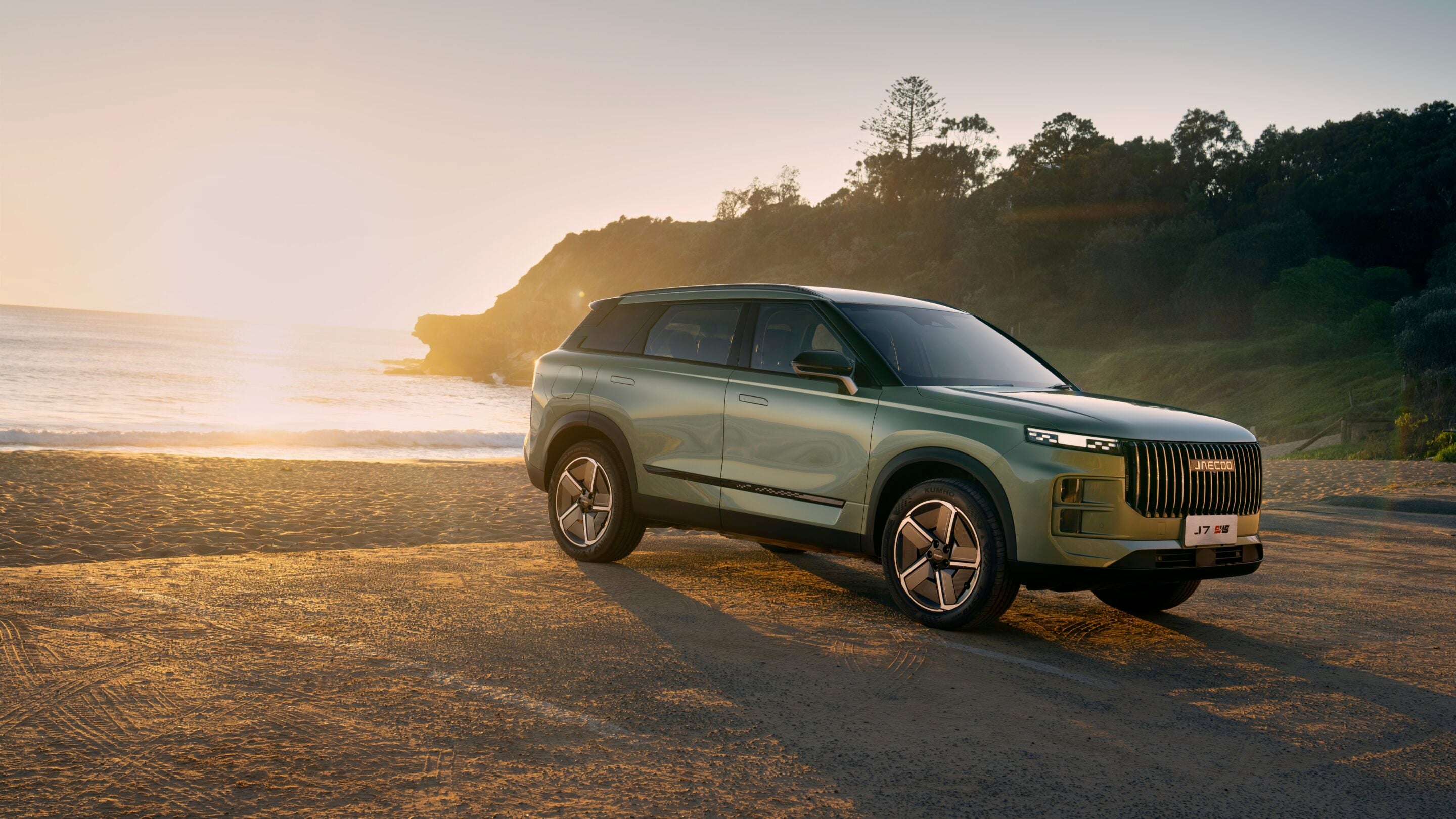 Green SUV parked on sandy beach at sunset with a rocky cliff and trees in the background