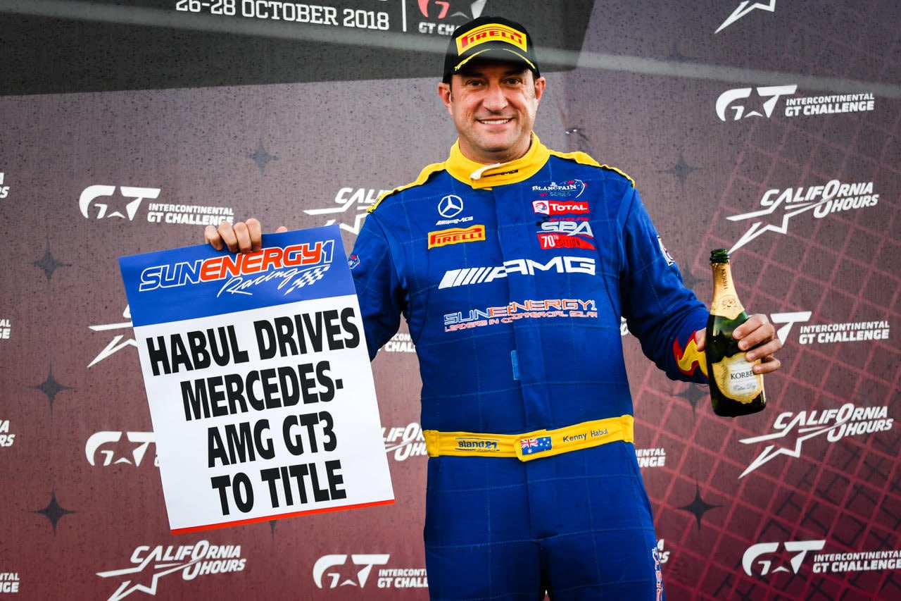 Kenny Habul on a podium in a blue AMG race suit, holding a sign about Mercedes-AMG GT3 winning the title and a bottle of champagne.