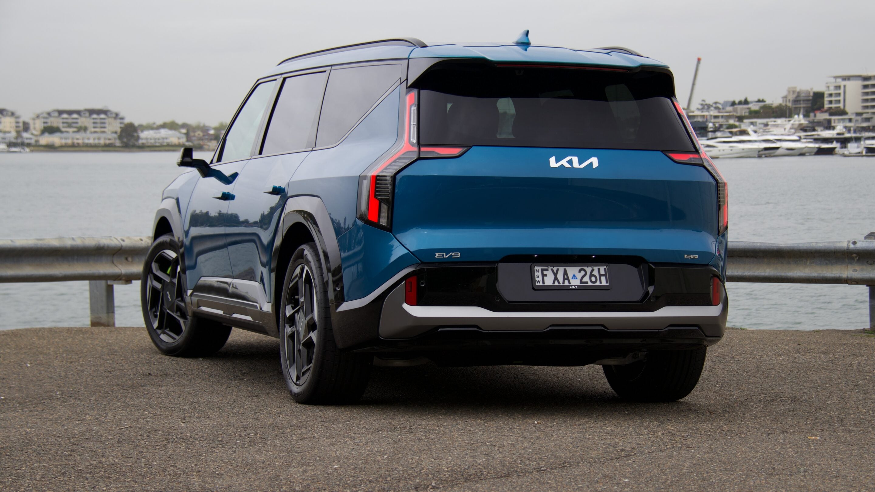 Blue Kia EV9 SUV parked by a waterfront marina, rear three-quarter view with boats and buildings in the background