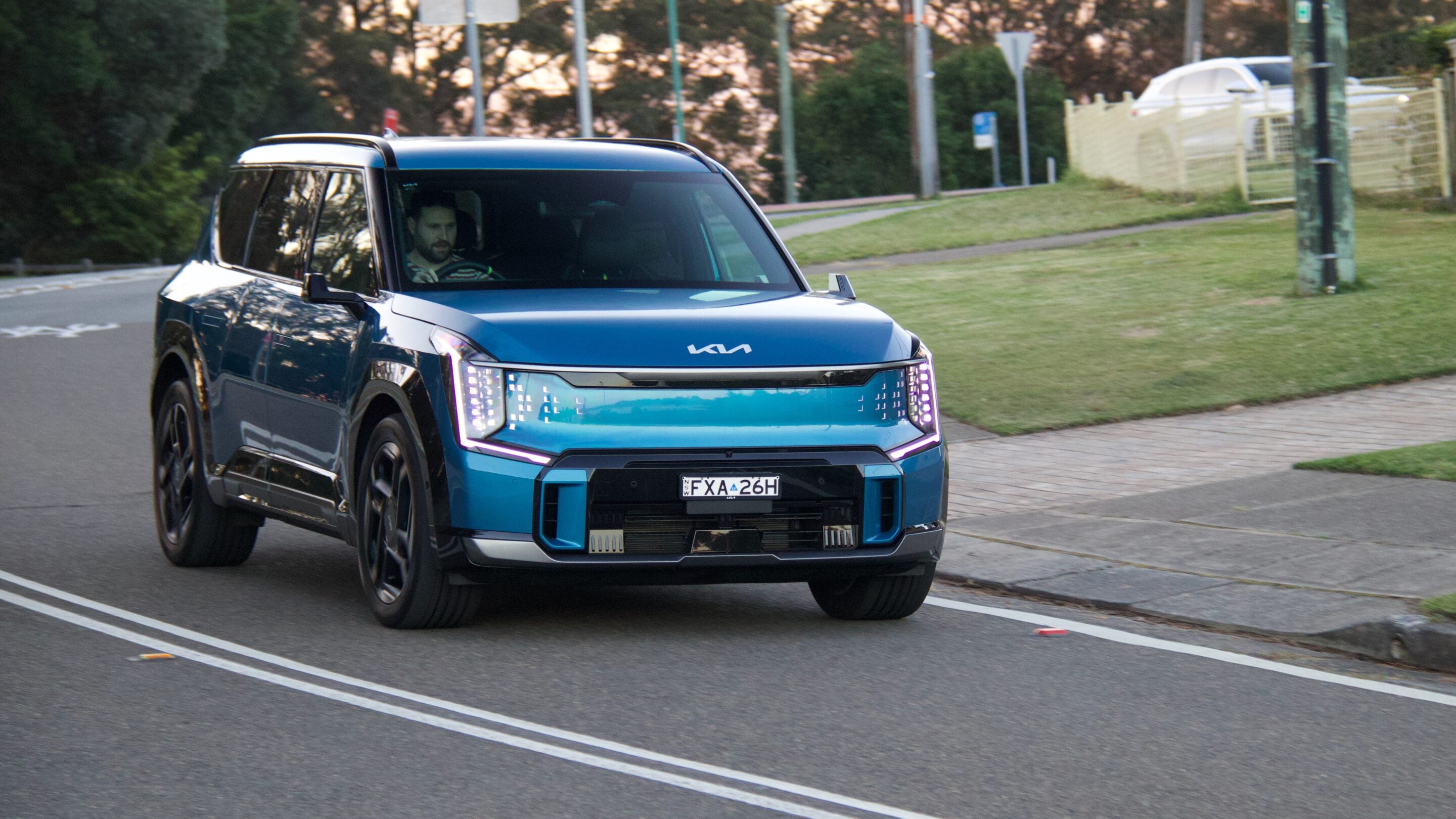 Blue SUV driving on a city street, driver visible through the windshield, trees and fence in the background.