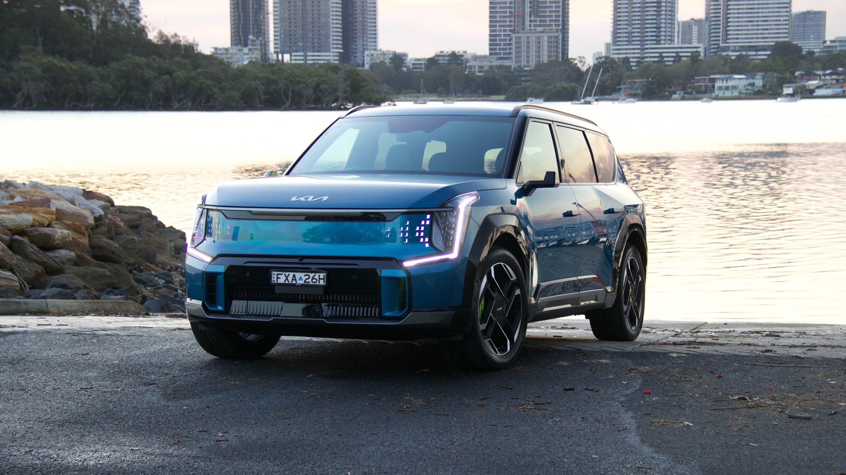 Blue modern SUV parked along a rocky waterfront with a city skyline in the background.