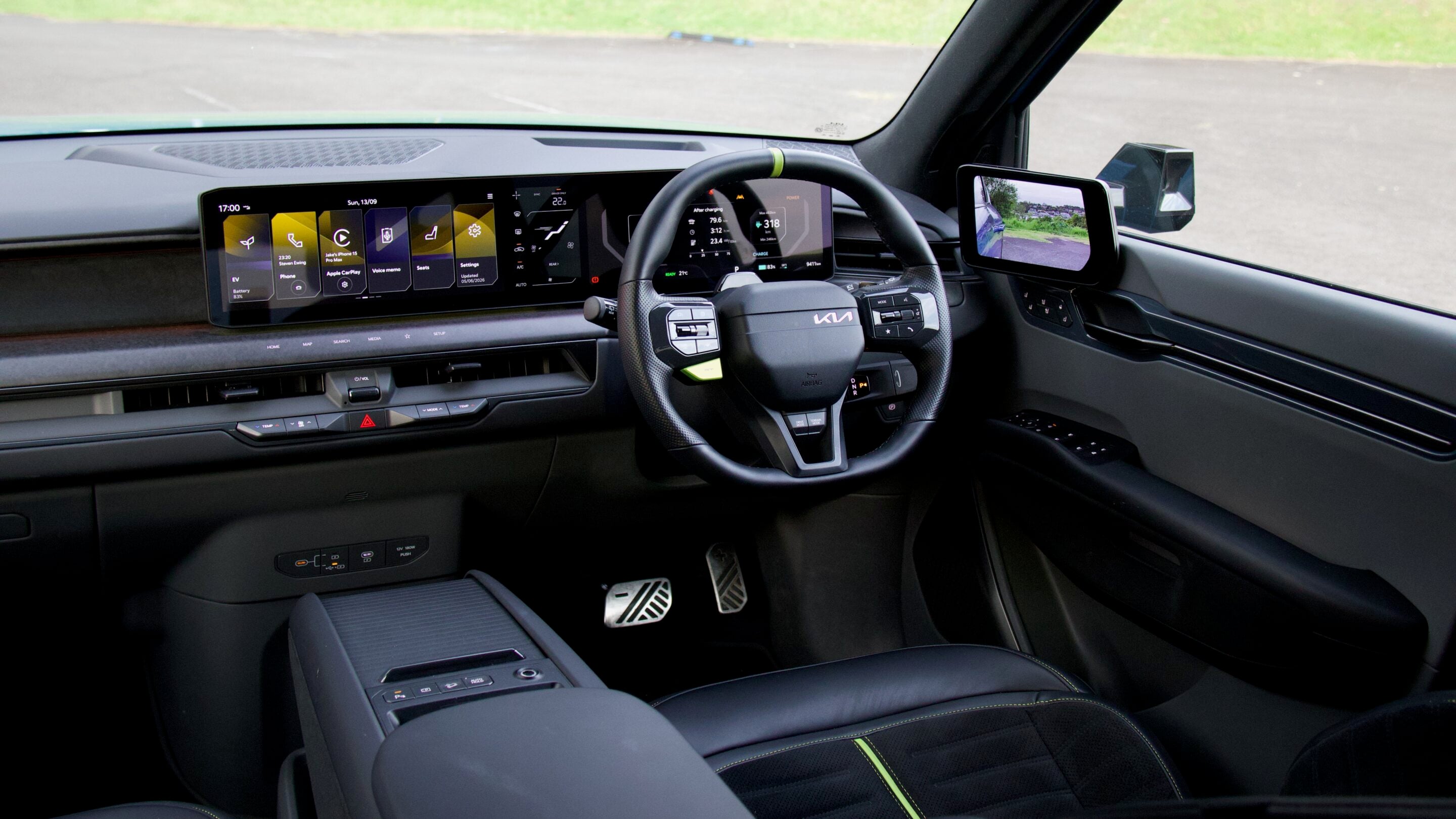 Modern car interior with a wide digital dashboard spanning the windshield and a sporty steering wheel. Large touchscreen displays and climate controls visible, driver-side view through the window.