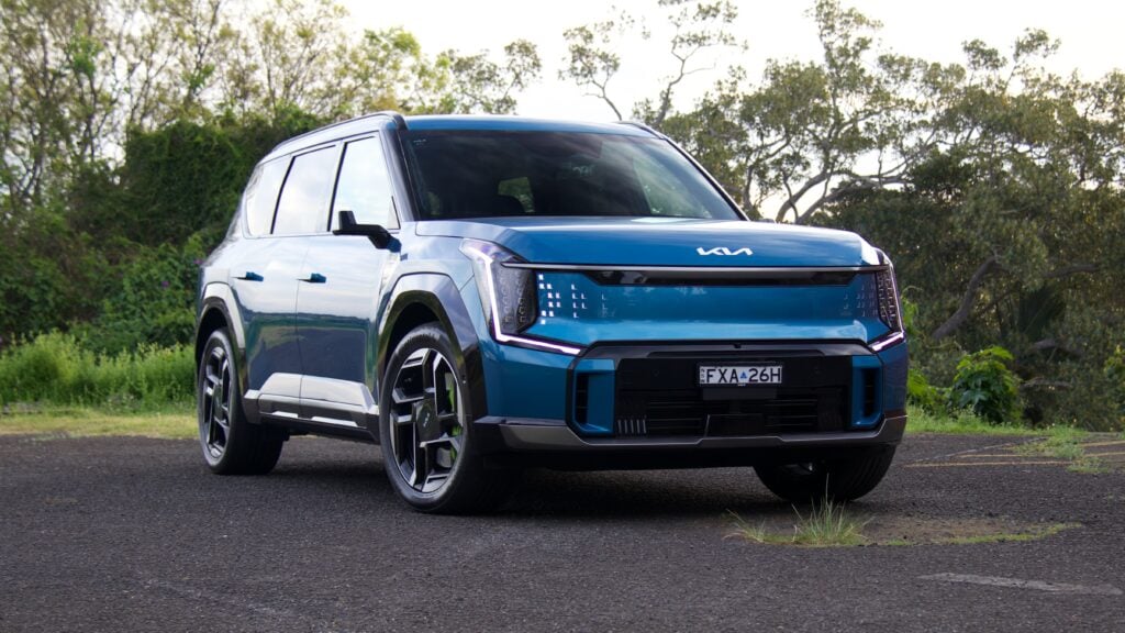 Front three-quarter view of a blue SUV parked on a paved lot with trees in the background, showing its bold grille and LED headlights.