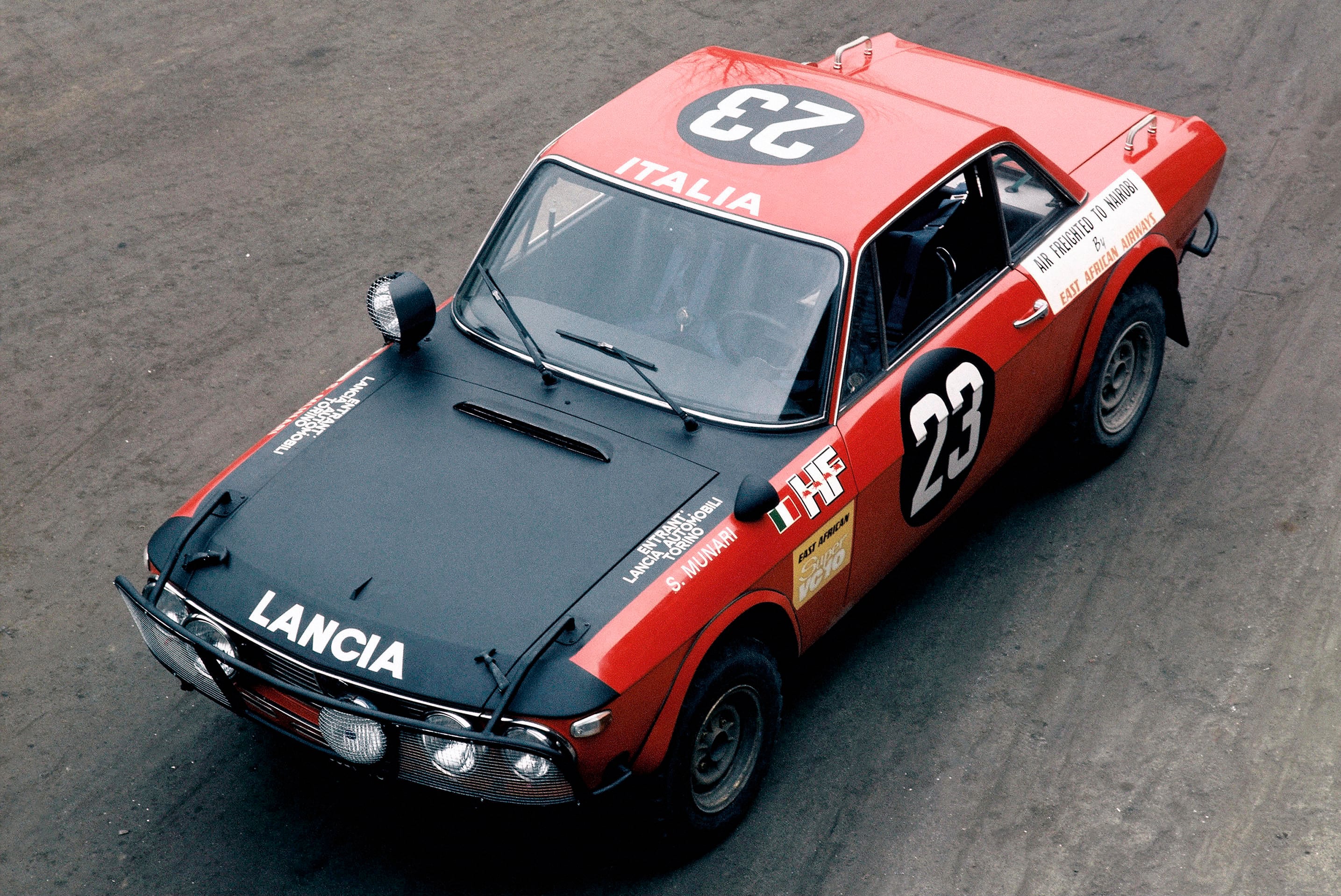 Top-down view of a red Lancia race car with number 23 and 'Italia' on the hood, parked on asphalt.