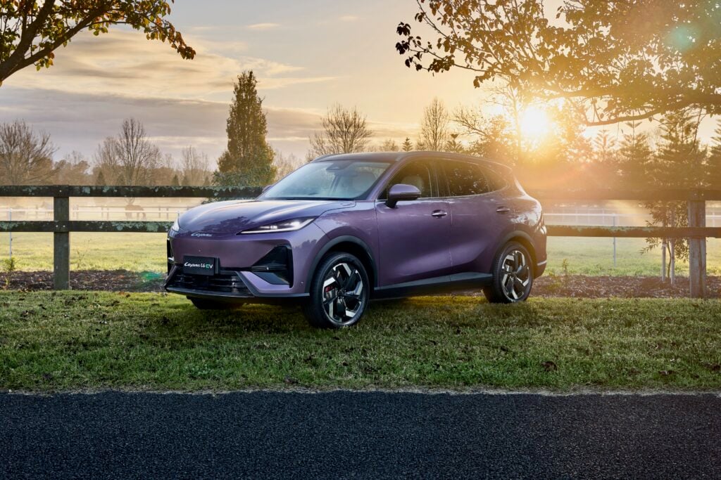Purple electric SUV parked on grass beside a wooden fence at sunset with trees in the background.