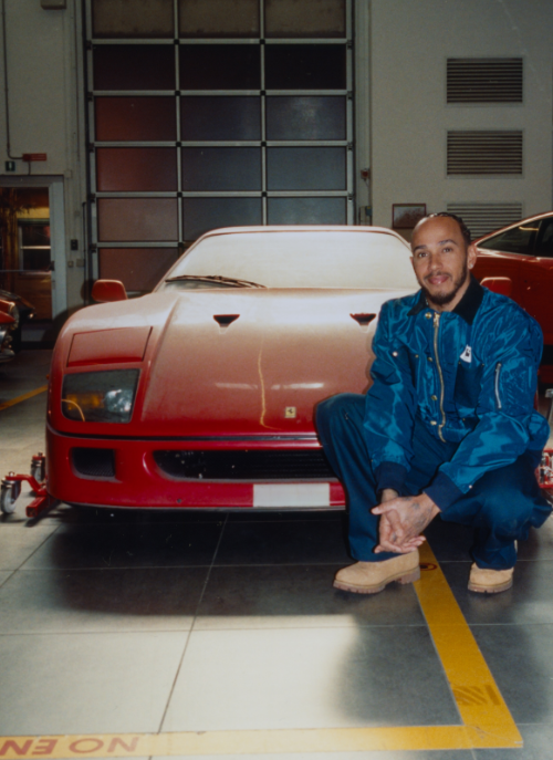 Man in a blue jumpsuit kneeling beside a red sports car in a garage or workshop environment.