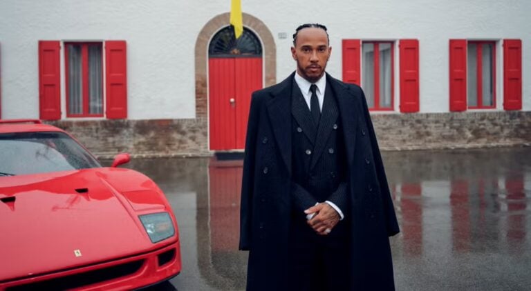 Man in a dark overcoat and suit stands beside a red sports car in front of a white building with red shutters.