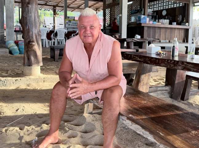 Man in a pink sleeveless shirt sits on a wooden bench on a sandy beach under a shaded, open-air hut, looking at the camera with hands clasped.