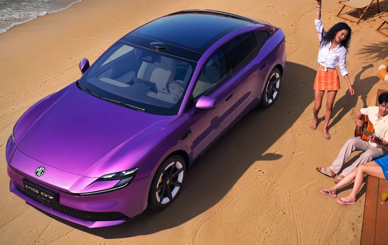 Purple MG car parked on a sandy beach with waves in the background; a group of friends nearby enjoying the day, one woman standing and another playing guitar nearby.