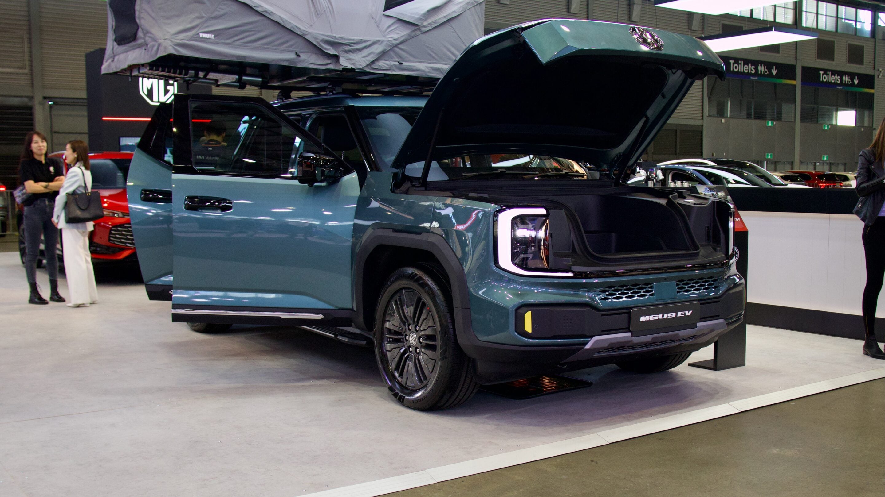 Blue MG MGUS EV electric SUV on display with open doors and raised rear hatch at an indoor auto show; people nearby.