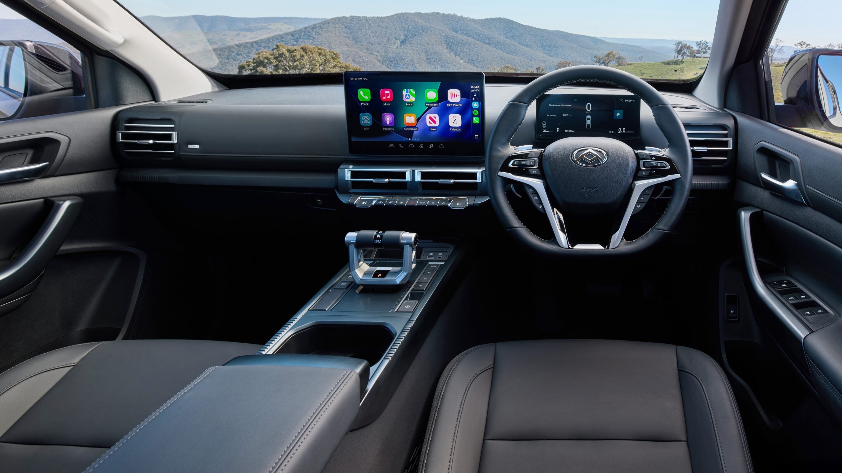 Wide view of a car interior showing a large central touchscreen, digital instrument cluster, and a futuristic gear selector in a premium cabin with gray leather seats.