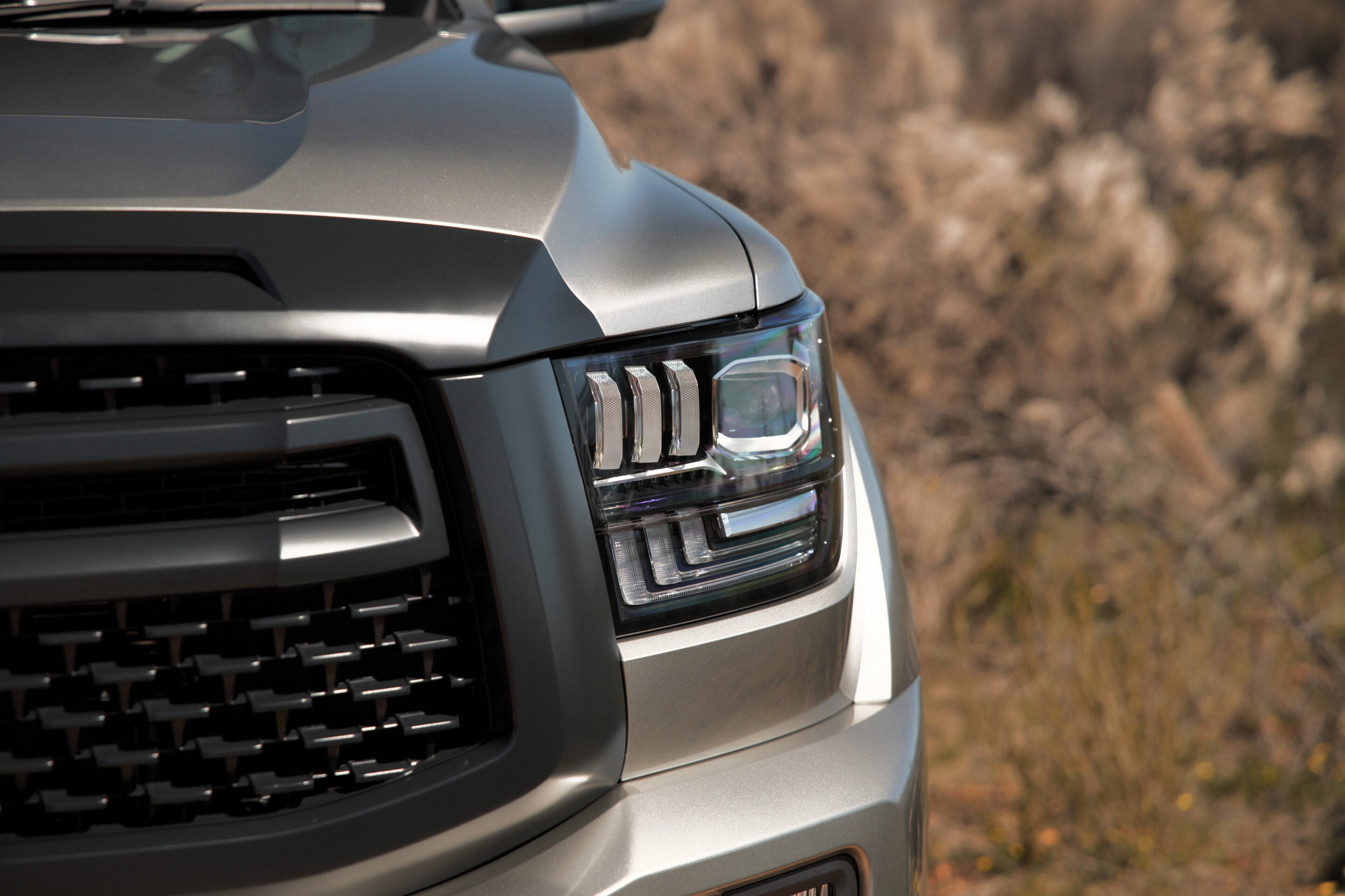 Close-up of the front left corner of a silver pickup truck, showing the headlight, hood crease, and grille with a blurred natural background.