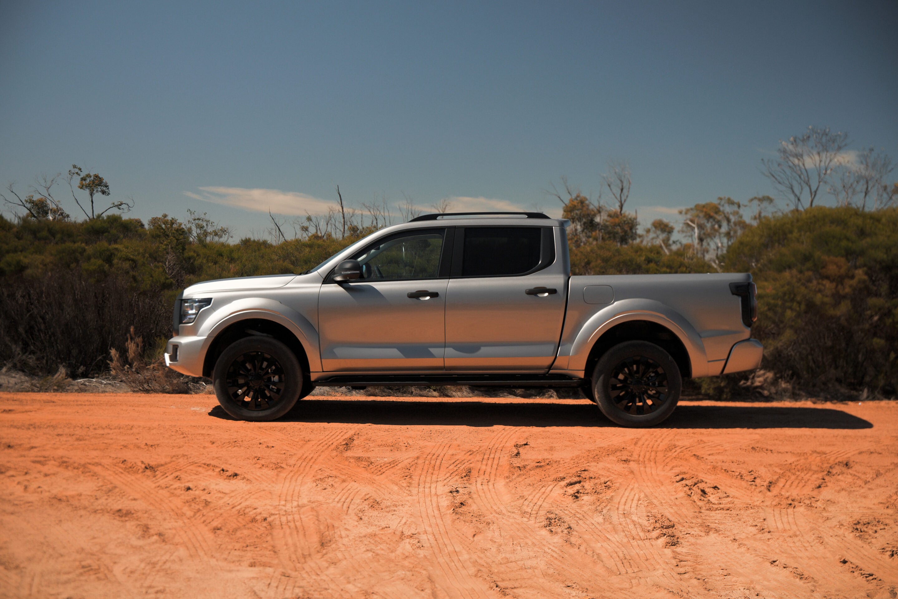Side view of a silver pickup truck parked on an orange dirt road in a desert landscape with scrubby bushes under a clear blue sky