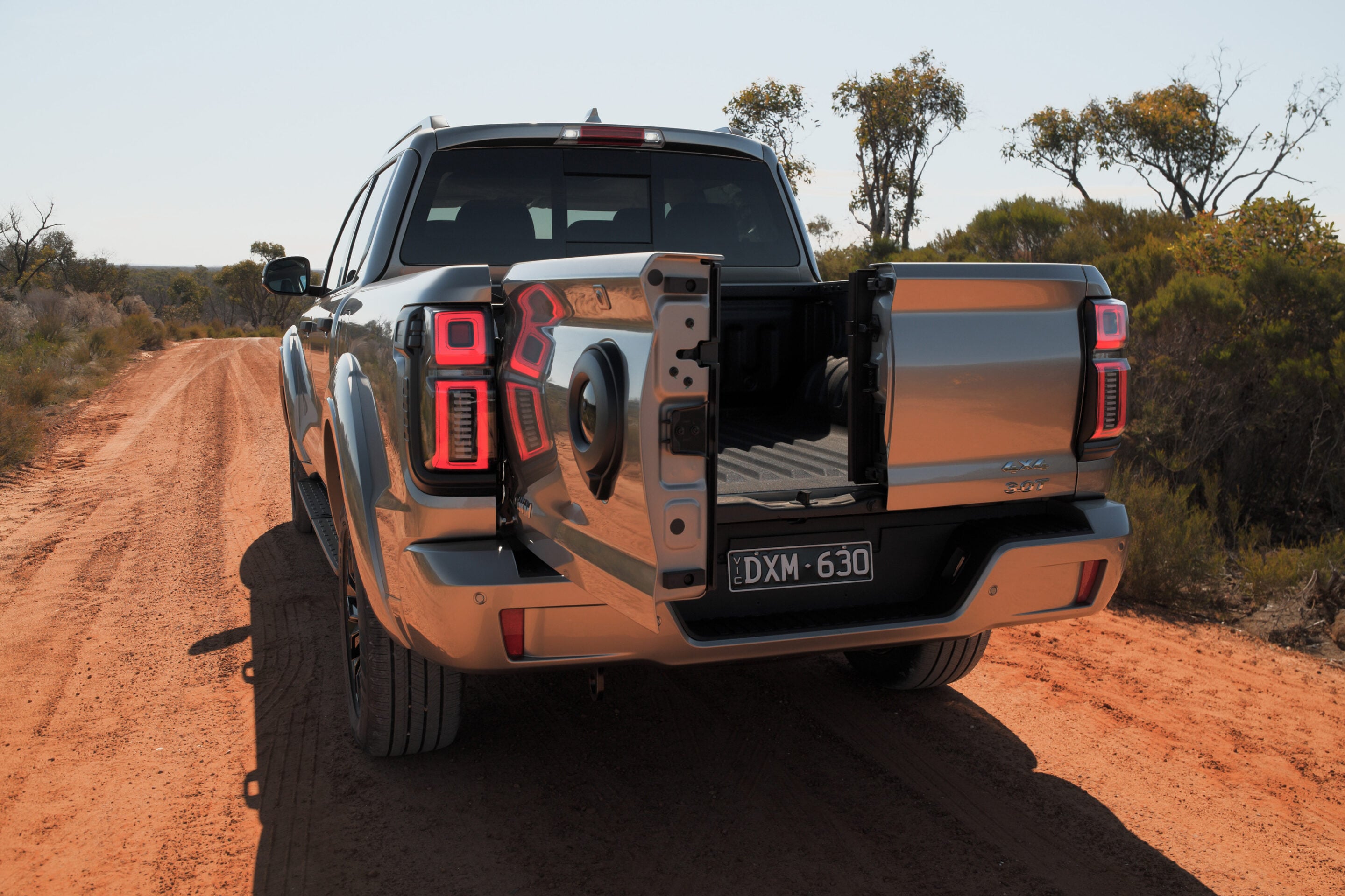 Silver pickup truck with bed open on a dusty dirt road in a dry, rural landscape at sunset.