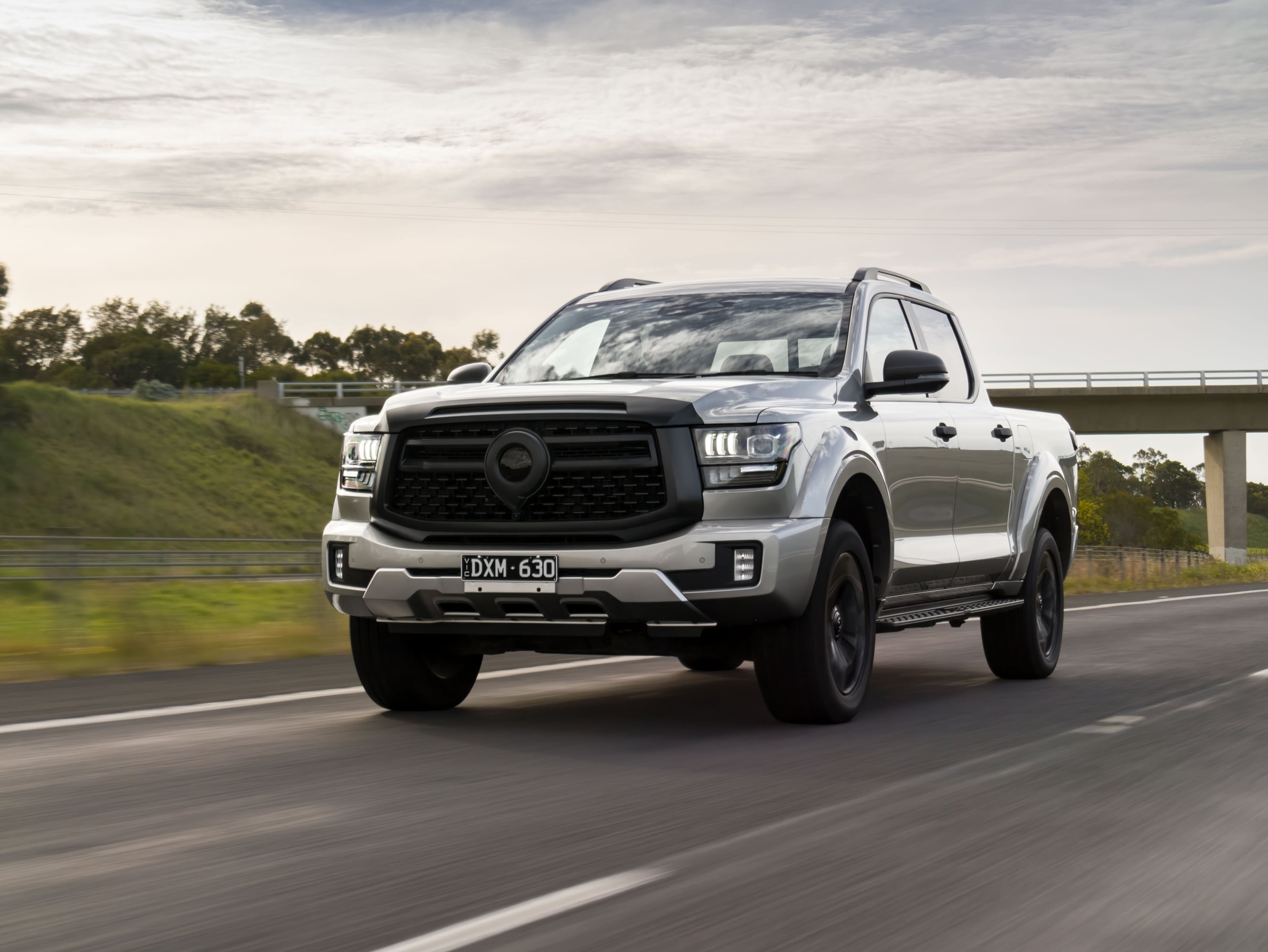 Silver pickup truck driving on a highway, front three-quarter view with a bridge and cloudy sky in the background.