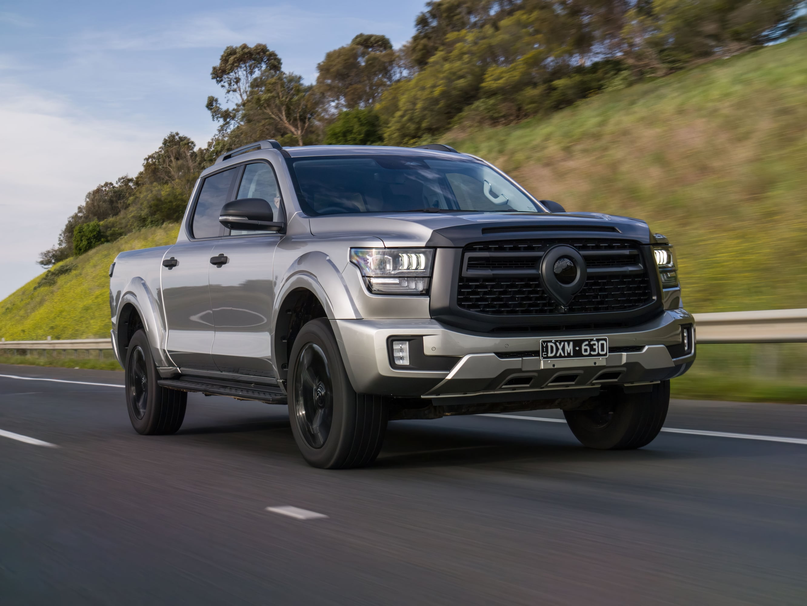 Front three-quarter view of a silver pickup truck driving on a highway with blurred green hills in the background.