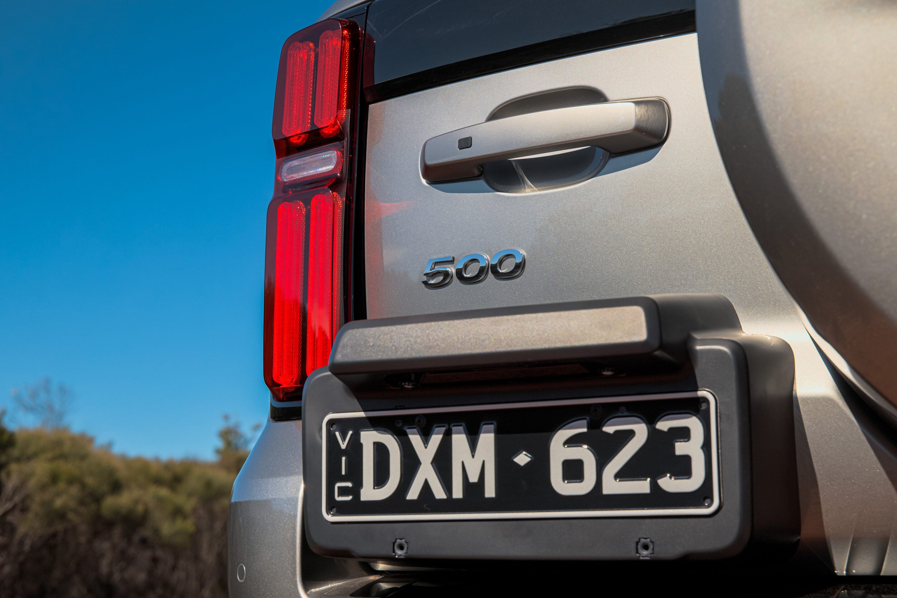 Rear view of a silver SUV showing a tall red tail light, tailgate handle, and a license plate visible with characters.