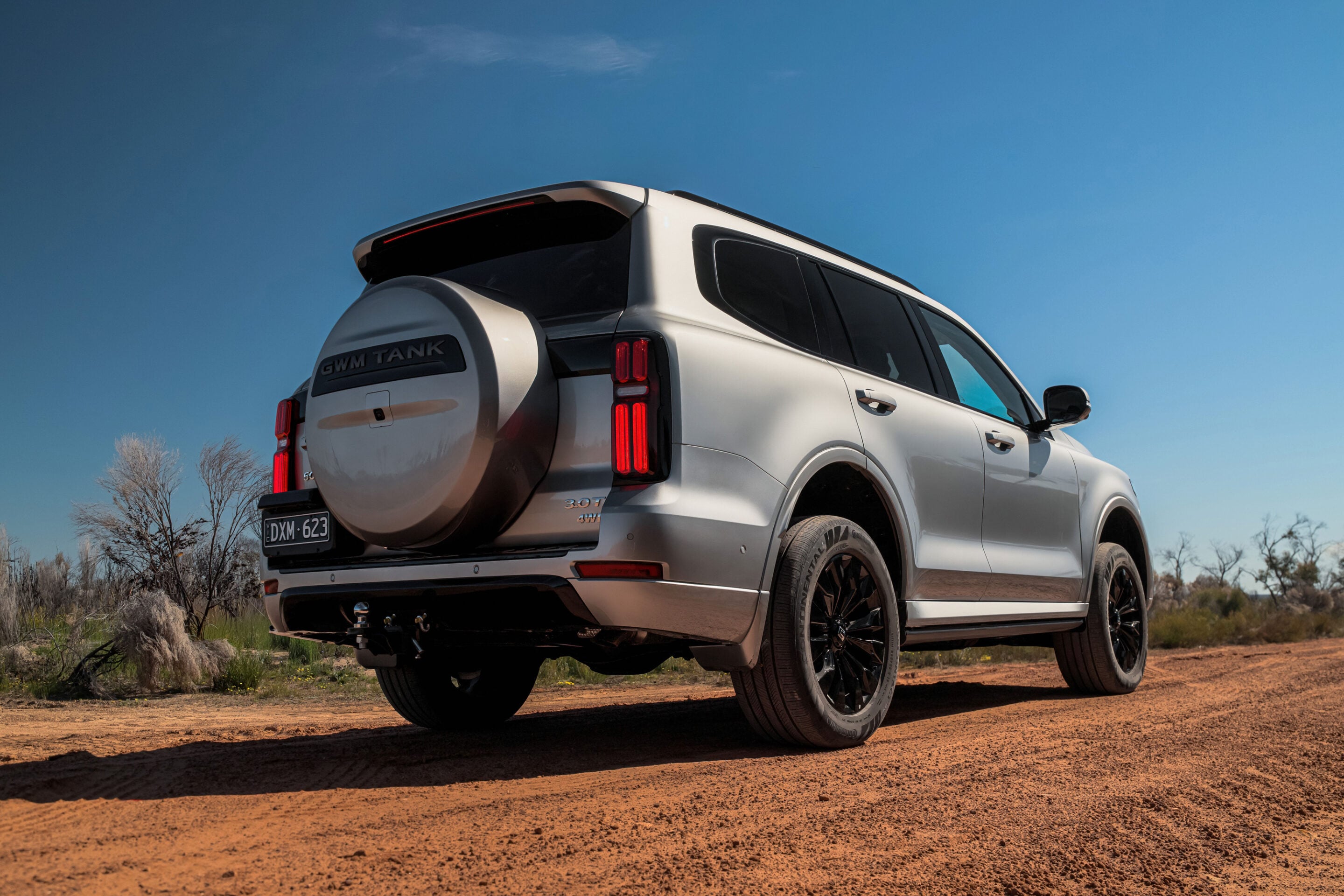 Rear three-quarter view of a white SUV with a spare-tire on the back, parked on a dirt road under a clear blue sky.