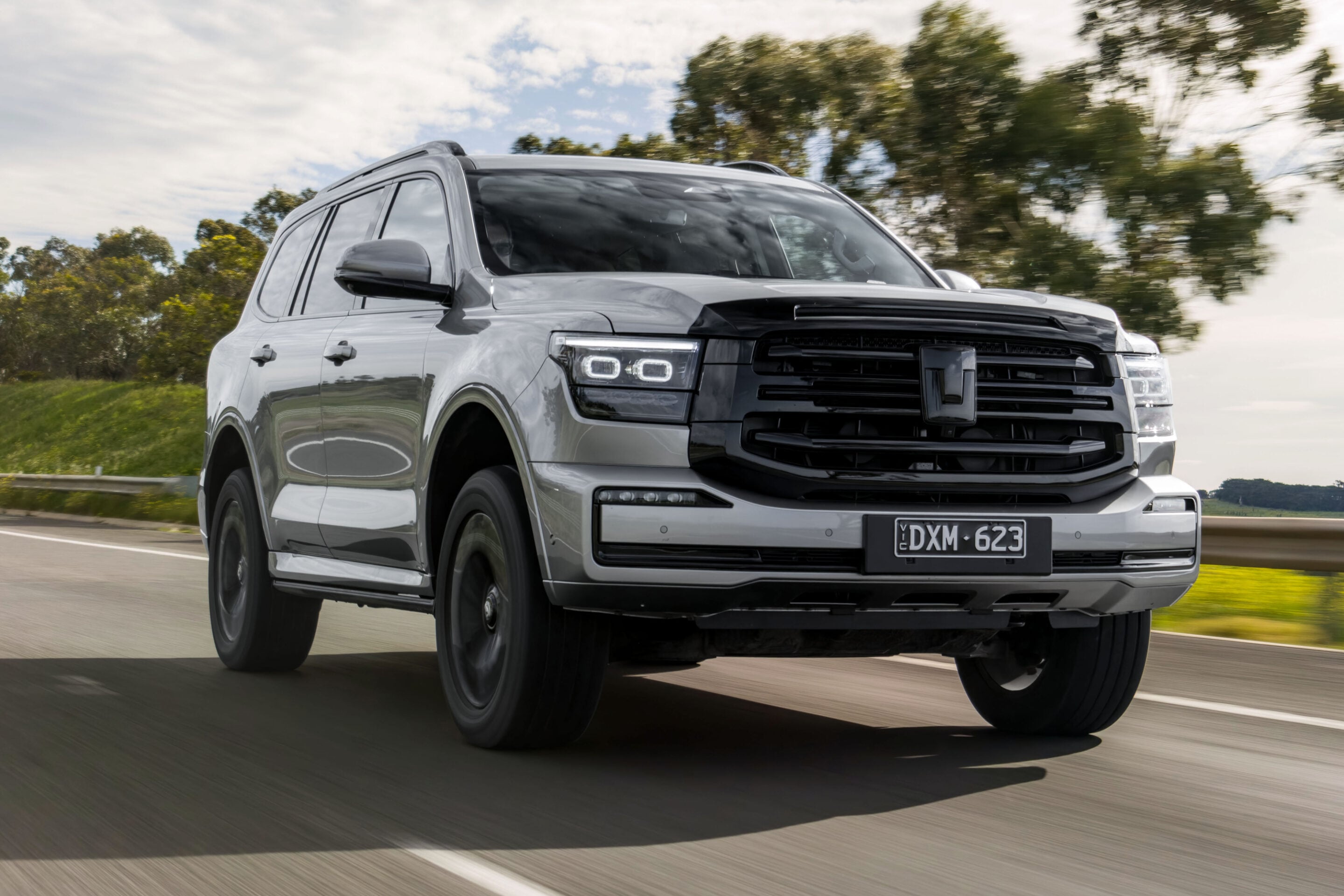 Silver SUV driving at speed on a highway with trees in the background, front grille and headlights visible.