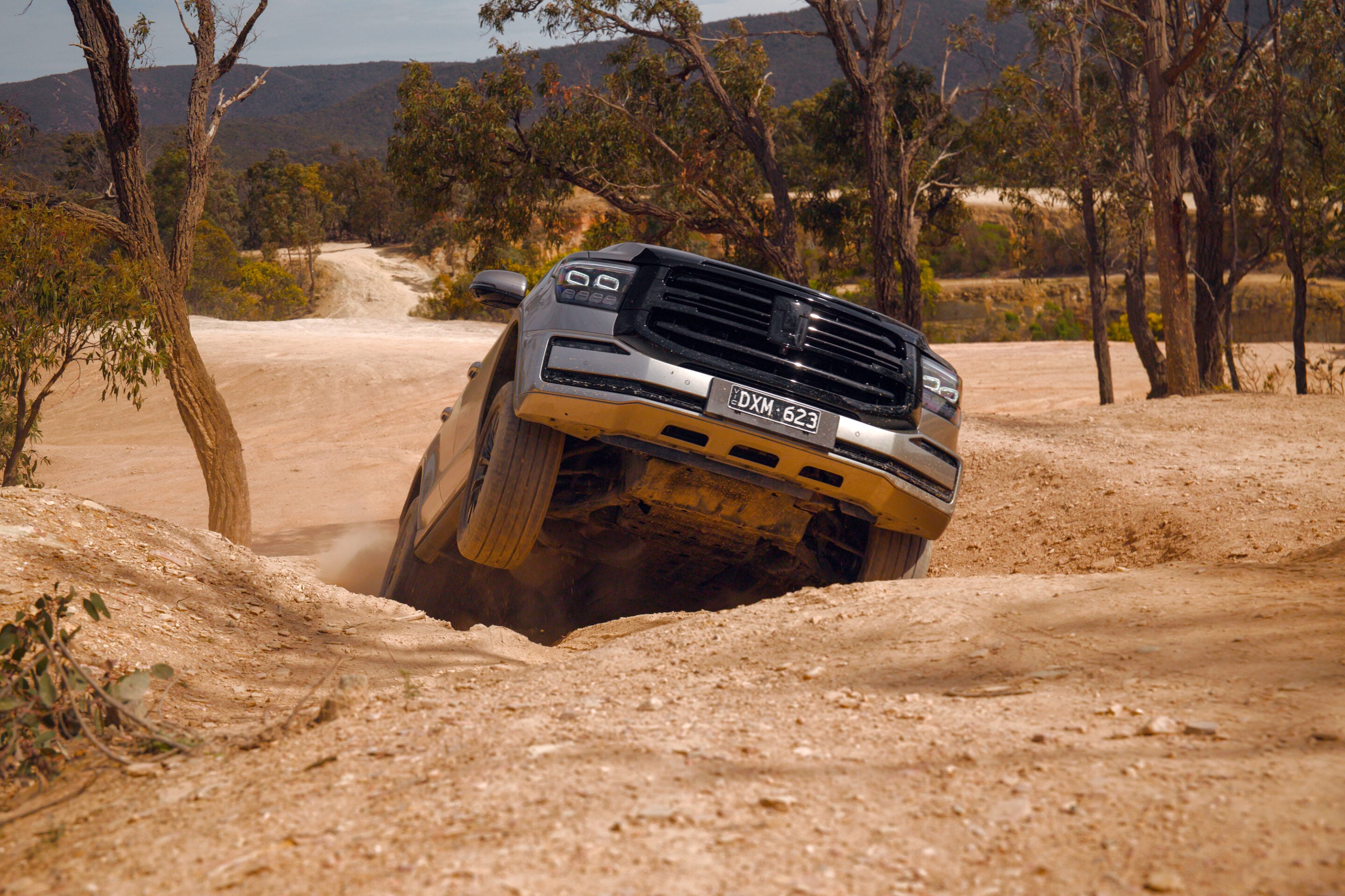Dark SUV tilting on a dusty, rutted dirt trail with dry, sparse trees and hills in the background.
