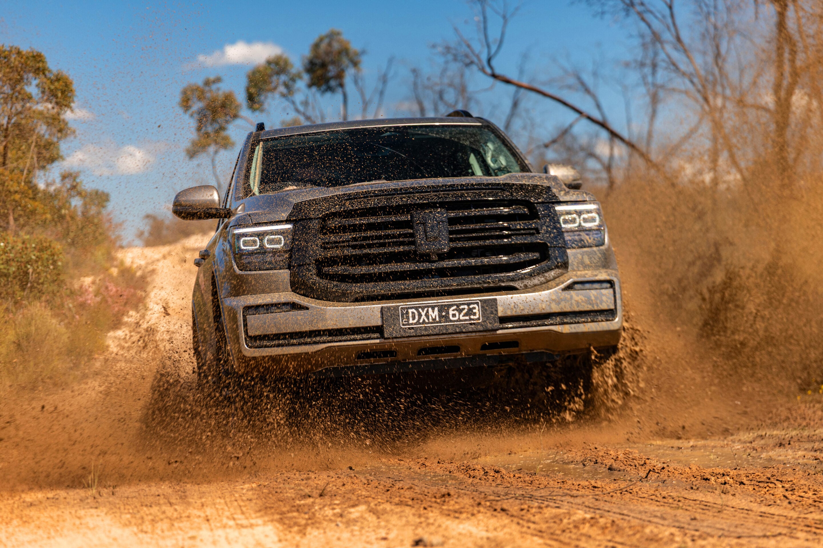 Front view of a mud-splattered pickup truck driving at speed on a dirt track, kicking up a large cloud of dust.