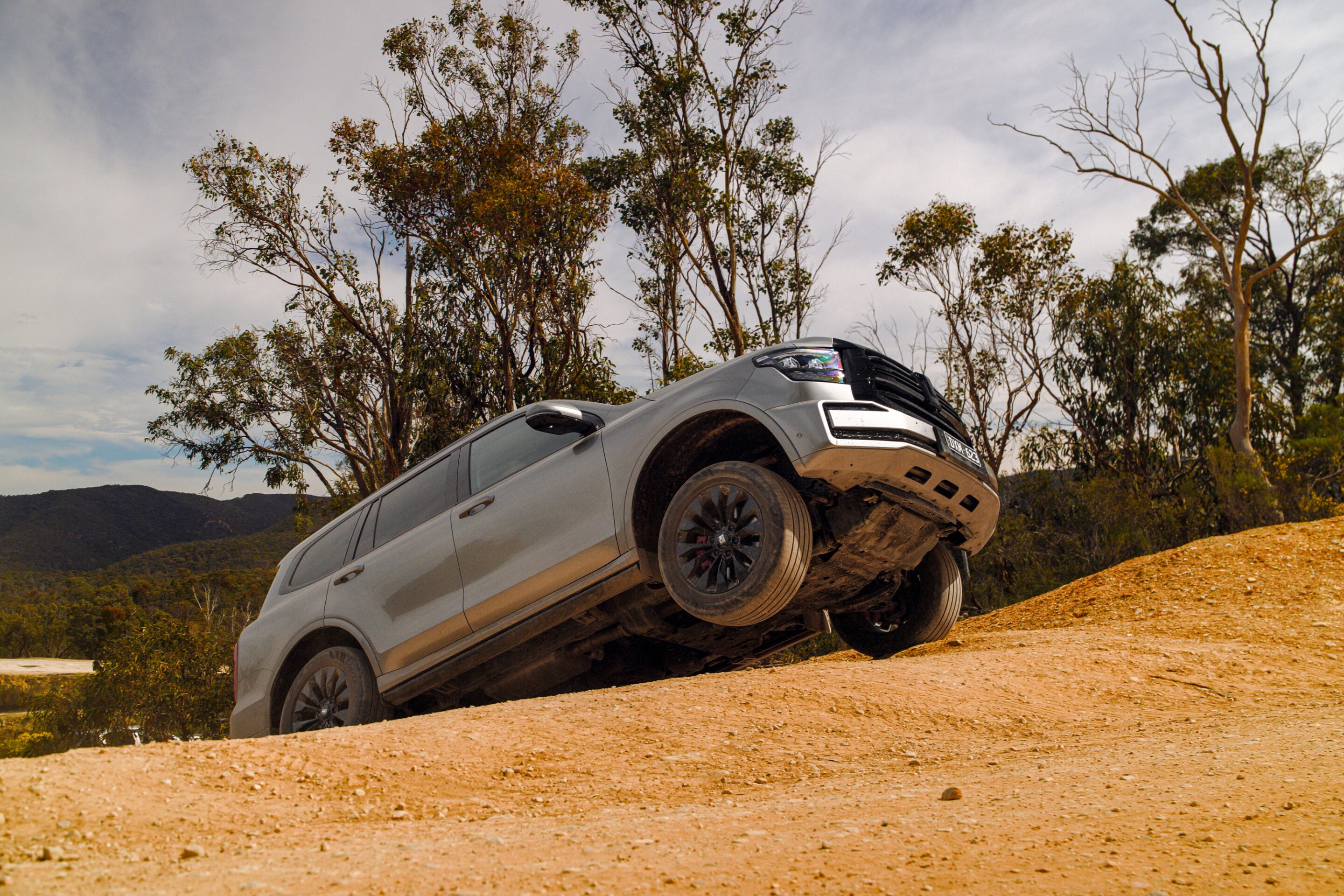 Off-road silver SUV tilting uphill on a dirt slope, wheels off the ground, with bare trees and hills in the background.