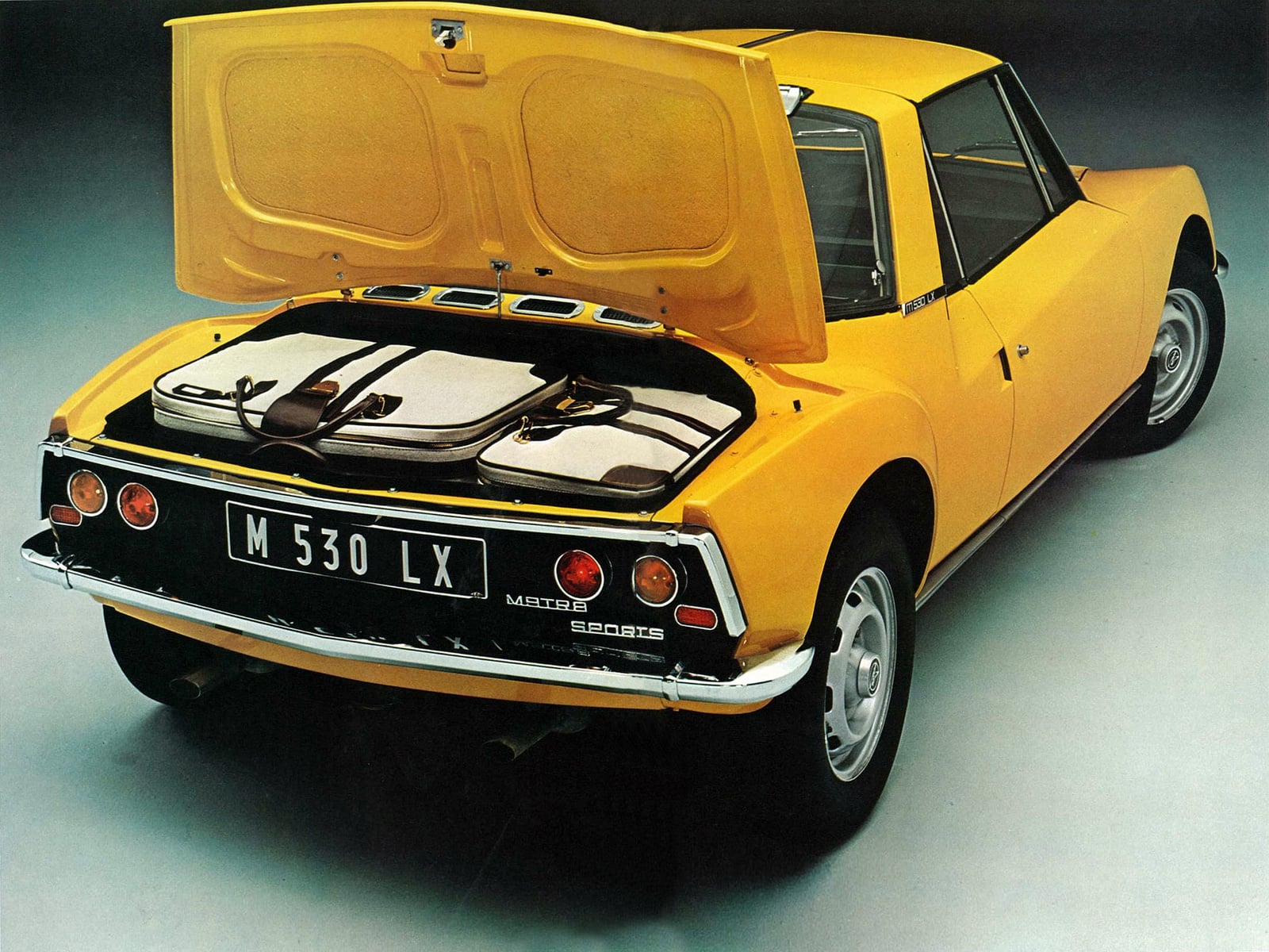Rear of a bright yellow vintage Matra sports car with the trunk open, revealing two white suitcases strapped inside the luggage area.