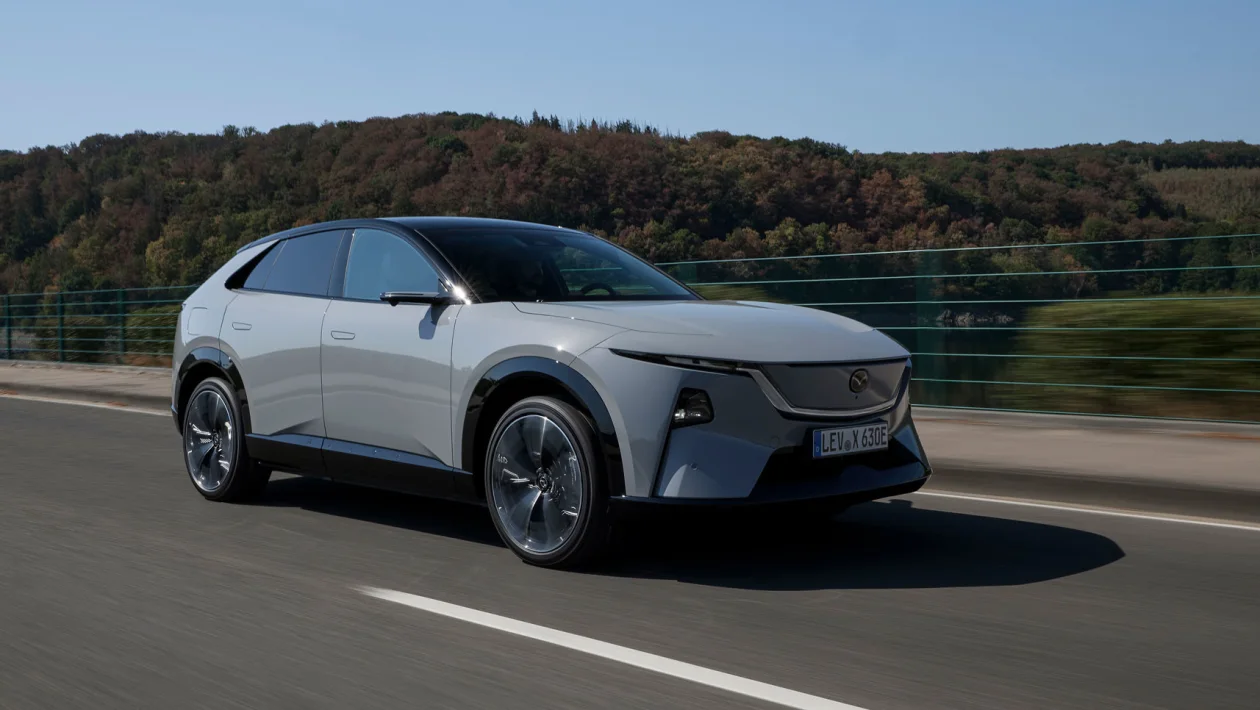 White electric crossover SUV driving on a highway with a forested hillside in the background.