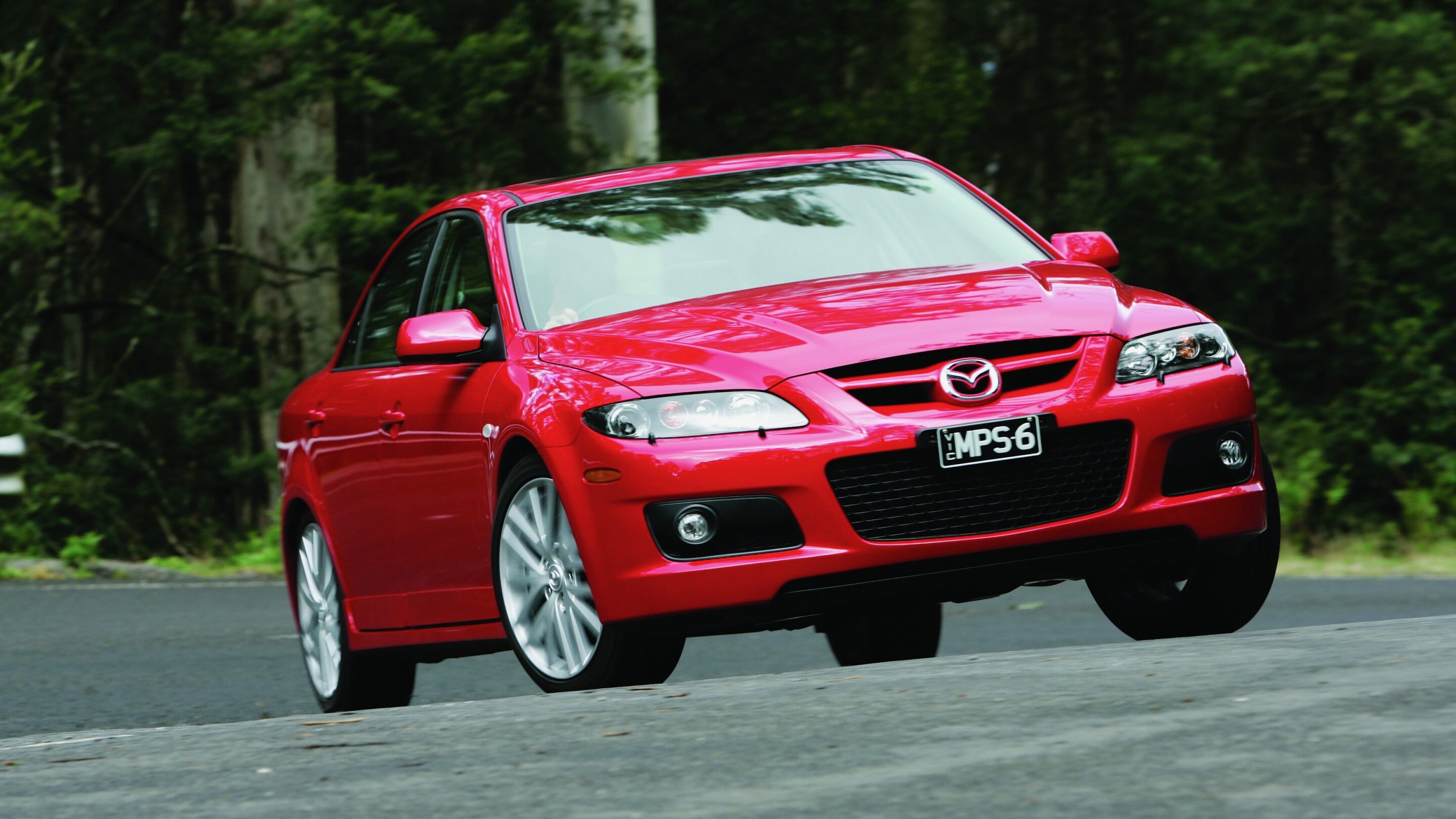Red Mazda sedan driving on a paved road with a forest background, angled toward the camera