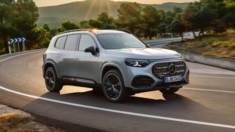 Silver Mercedes SUV driving on a winding mountain road at sunset, with trees and hills in the background.