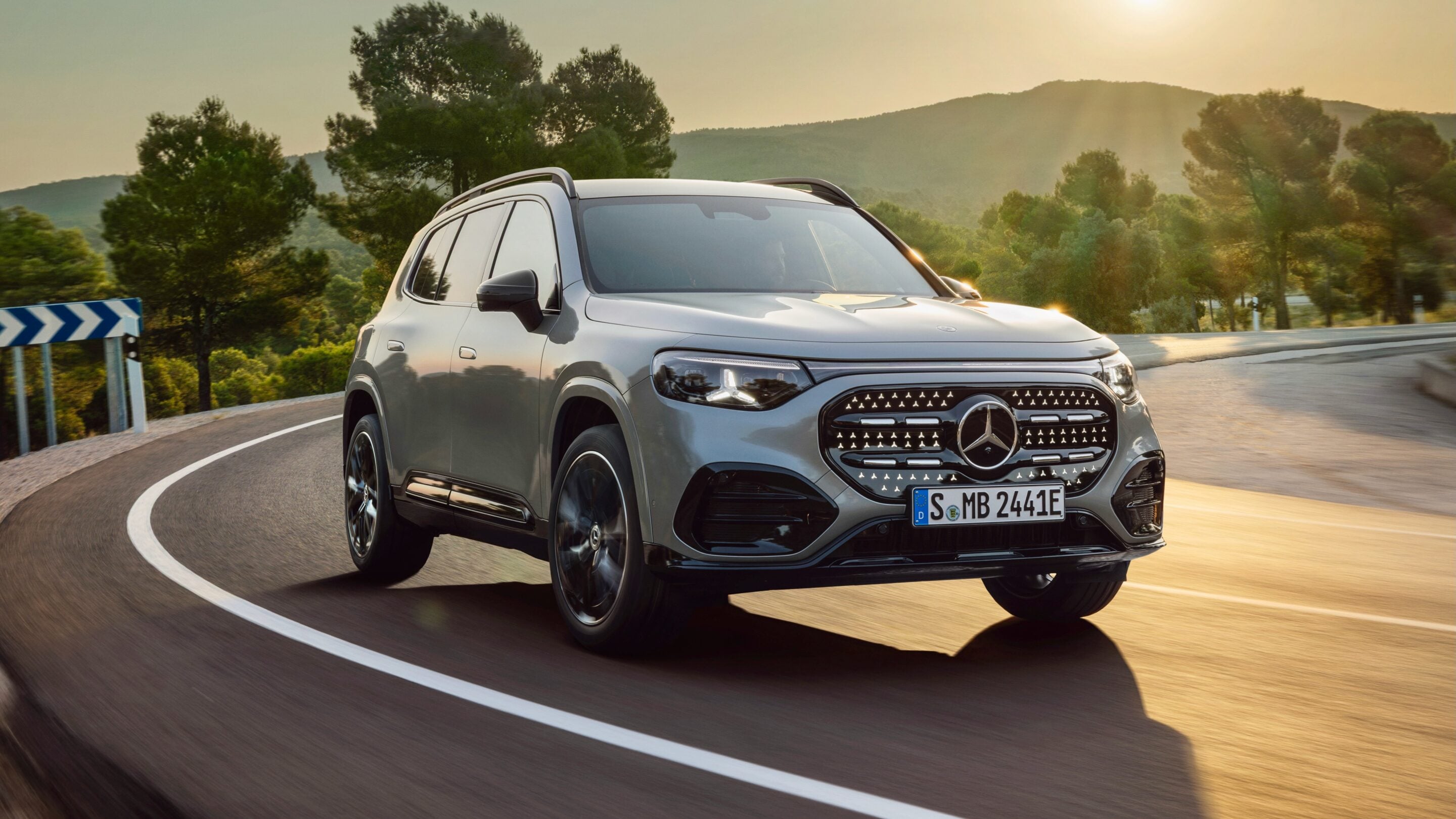 Silver Mercedes-Benz SUV driving on a winding mountain road at sunset.