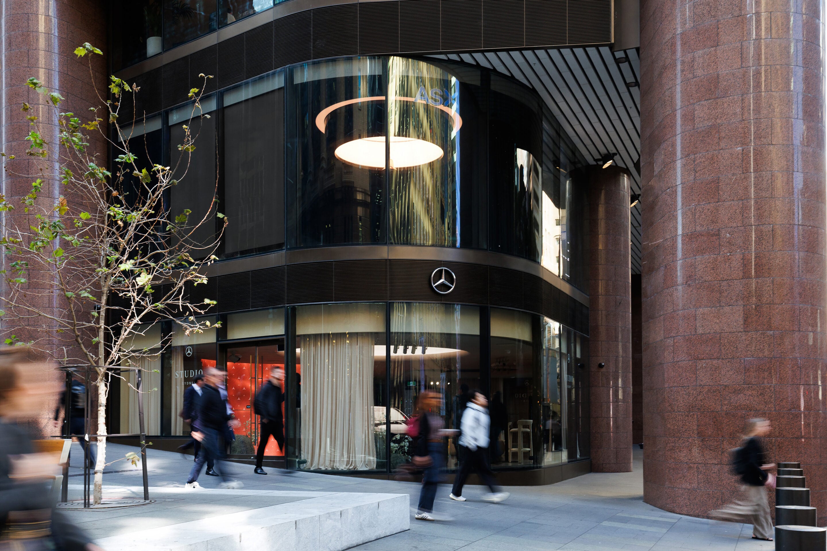 Glass-fronted Mercedes showroom on a curved façade with the logo visible, pedestrians walking by on a city street.
