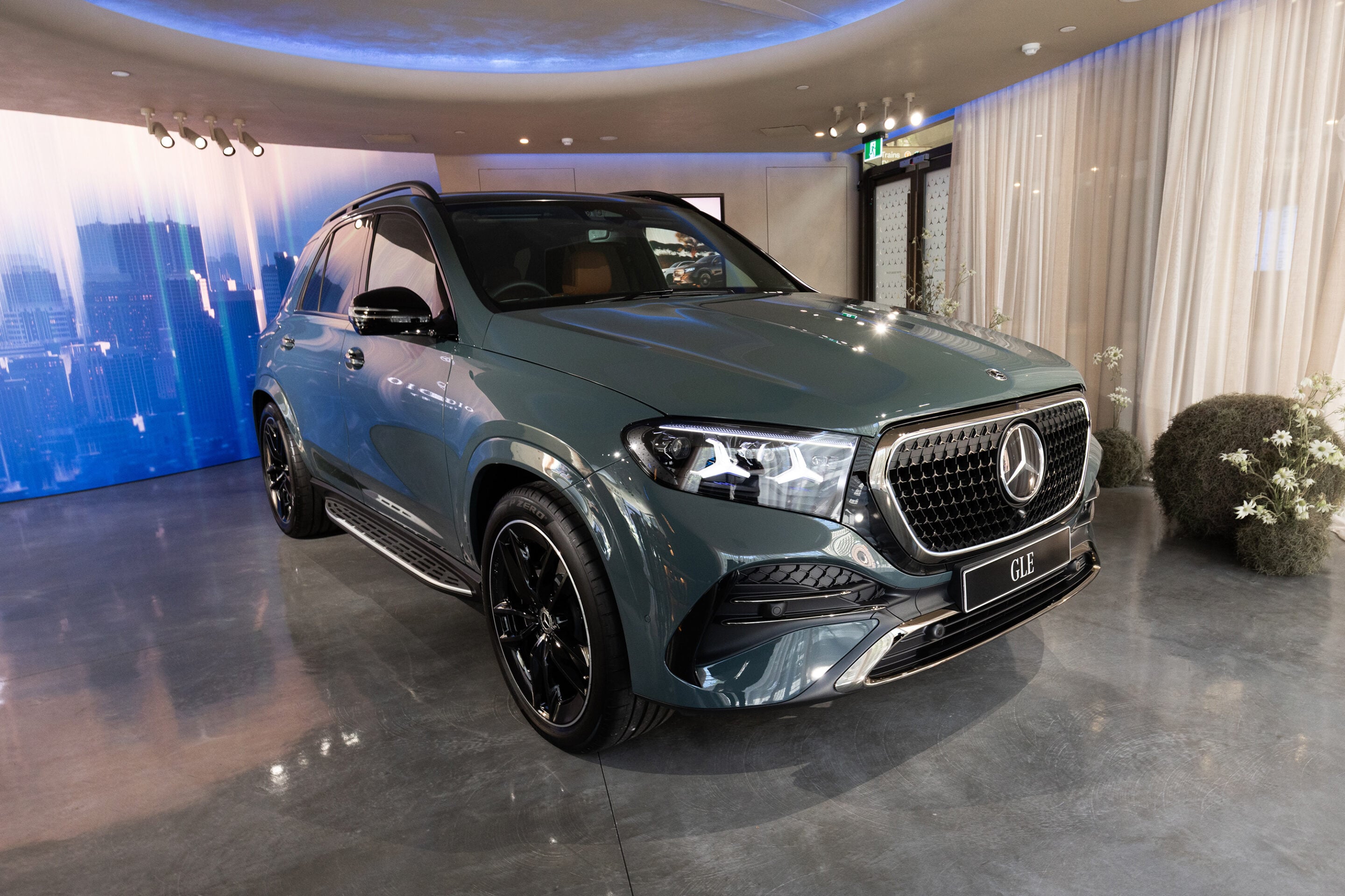 Blue-green Mercedes GLE SUV on display in a showroom with a cityscape backdrop and white curtains behind it