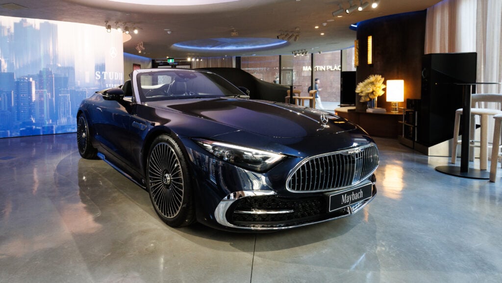Blue Maybach convertible on display in a showroom with a metallic grille and large chrome rims; cityscape backdrop and warm lighting in the background.