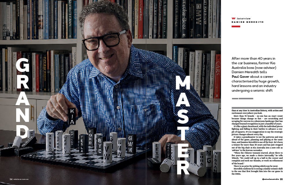 Smiling man in a blue patterned shirt plays a letter-tile chess game at a table, with a library backdrop; vertical words Grand and Master frame the scene for a magazine interview feature about his career.