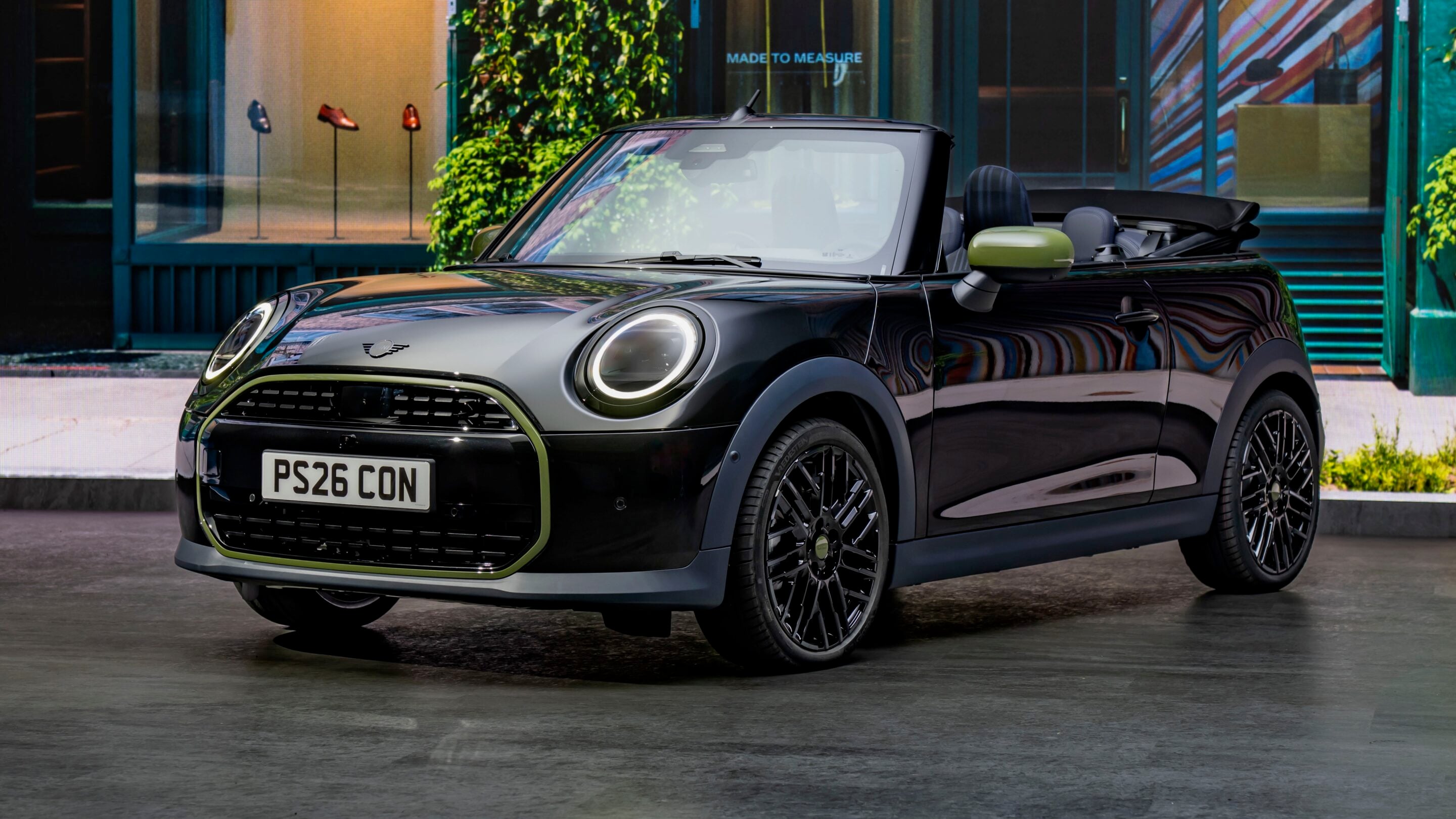 Black Mini Cooper Convertible with lime-green trim and alloy wheels parked outside a storefront; top down, license plate PS26 CON visible.