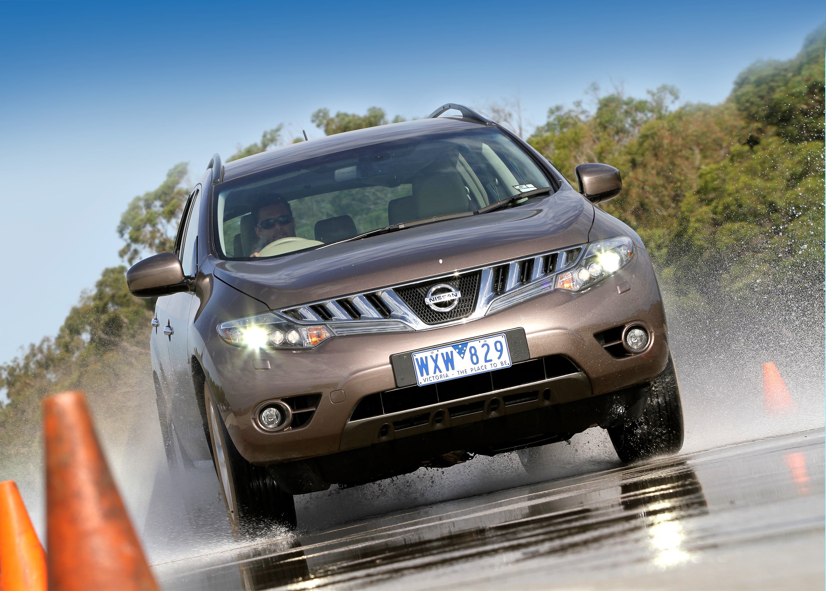 Front-three-quarter view of a brown Nissan SUV driving through a puddle, water spraying from the wheels on a wet road with orange traffic cones in the foreground and trees in the background.