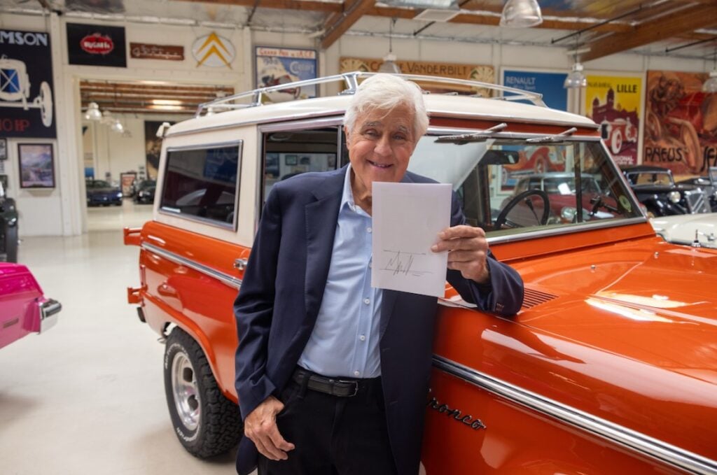 Smiling older man in a navy blazer holds a signed document beside an orange vintage van in a car showroom.