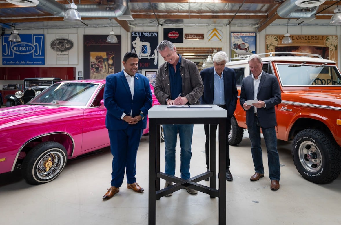 Four men in business attire stand around a signing desk in a vintage car showroom, with a pink coupe and an orange SUV in the background.