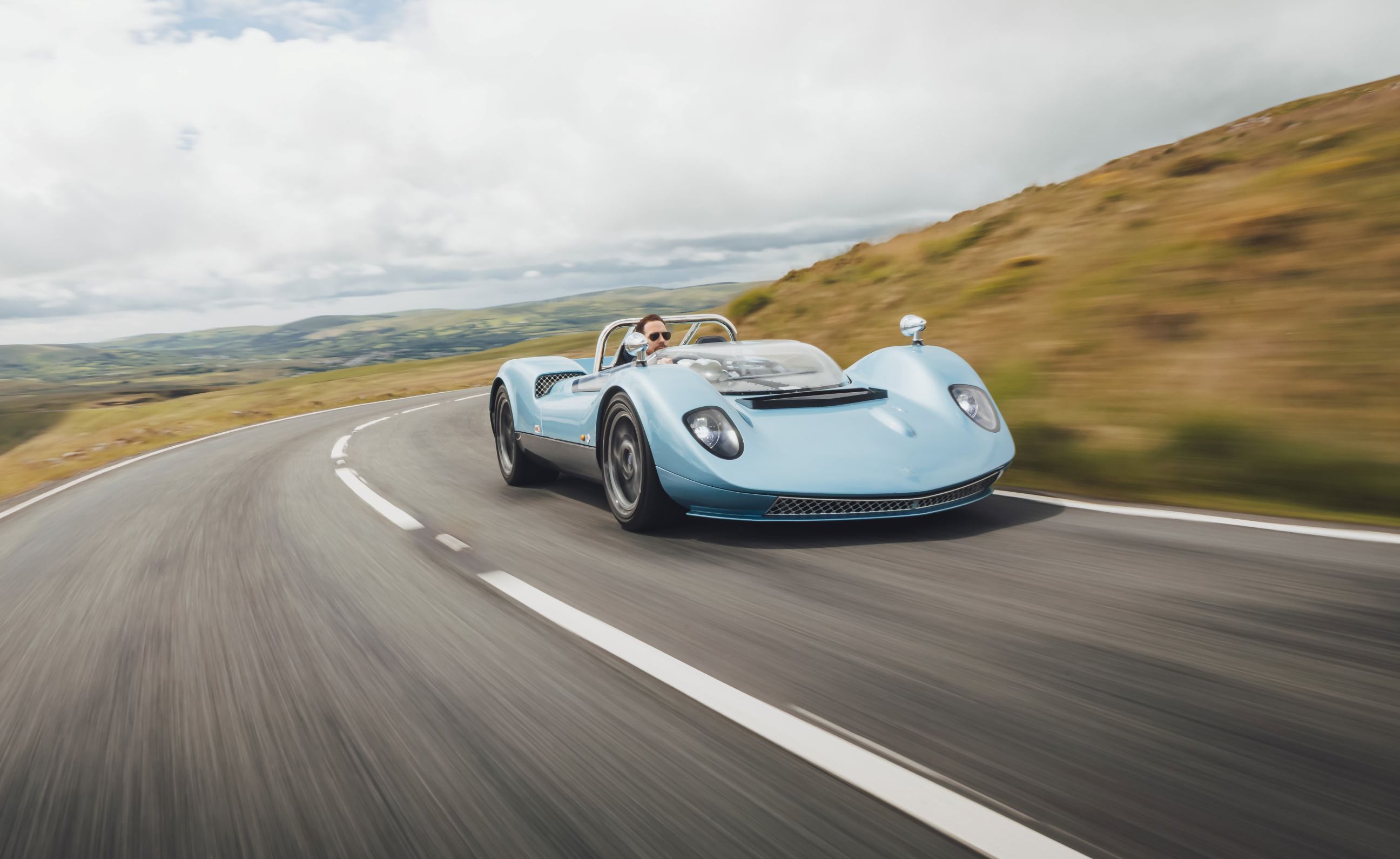 Blue vintage open-top sports car speeding along a winding countryside road with blurred scenery behind it.