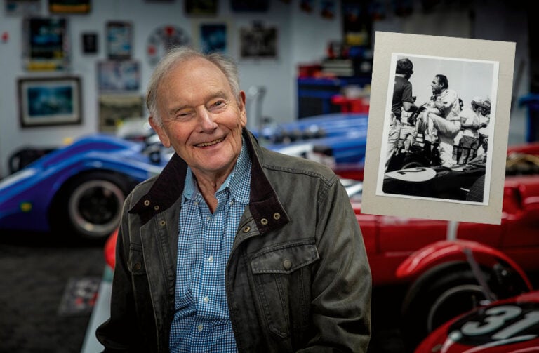 Smiling elderly man in a denim jacket sits in a garage filled with vintage race cars; a black-and-white photo is pinned nearby.