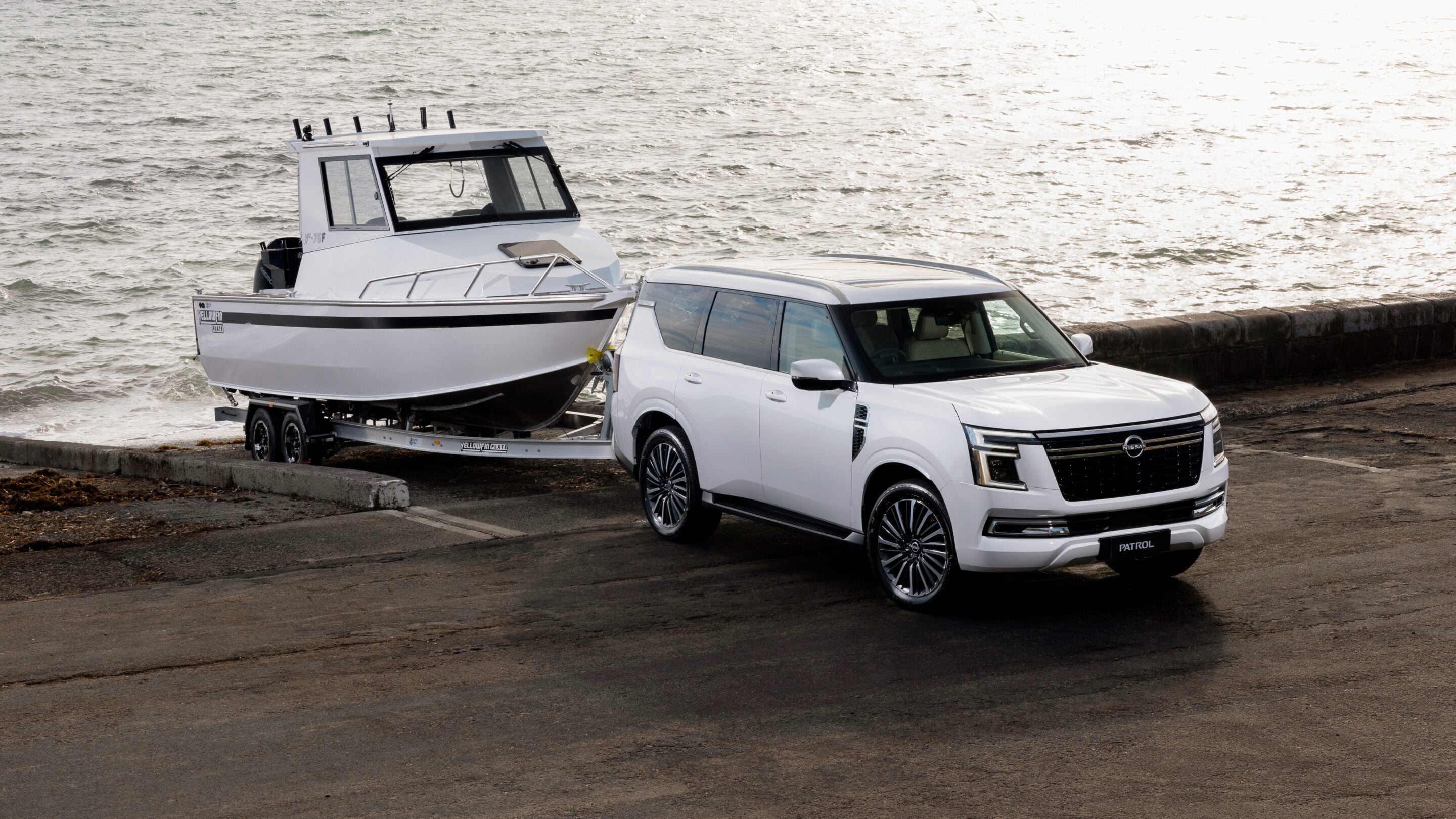 White Nissan Patrol SUV towing a white motorboat on a trailer along a seawall by the water.