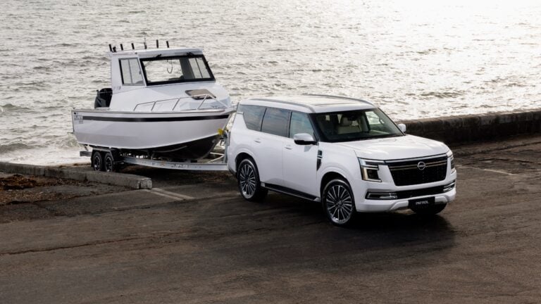 White Nissan Patrol SUV towing a white motorboat on a trailer along a seawall by the water.